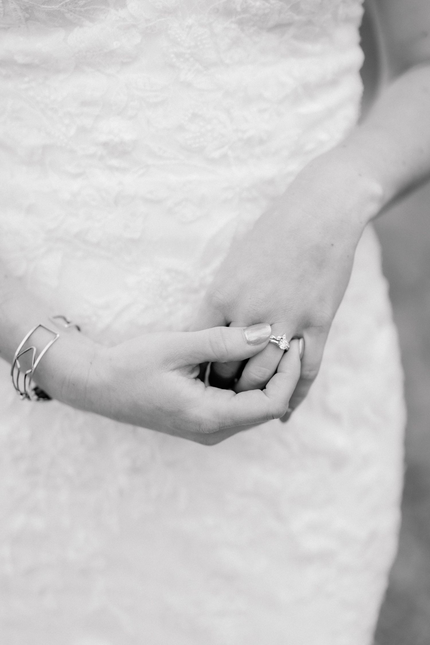 Black and white photo of a bride in lace dress clasping hands showing engagement ring and delicate bracelets.