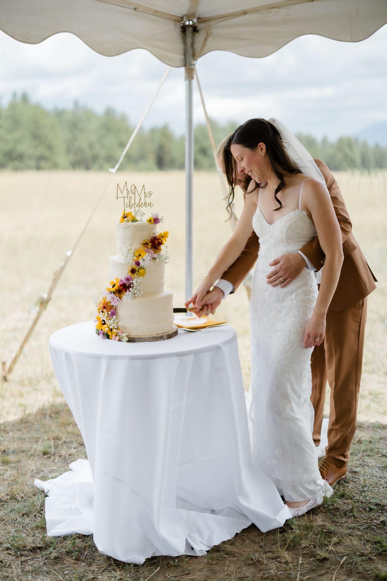 Bride and groom cutting a three-tier white wedding cake decorated with colorful flowers outdoors under a tent.