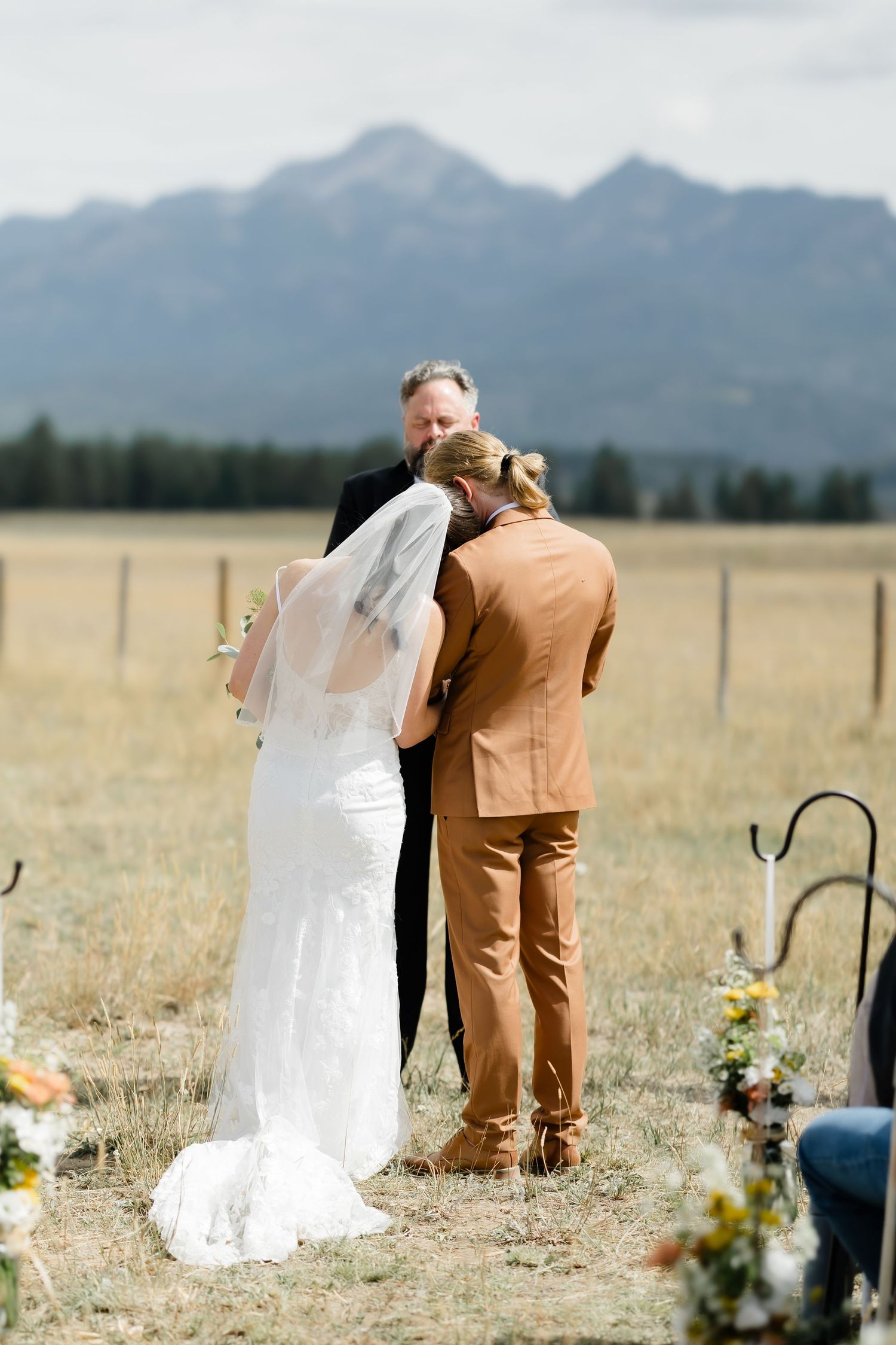 Outdoor wedding ceremony in a mountain meadow with bride in white gown, groom in tan suit, and officiant.