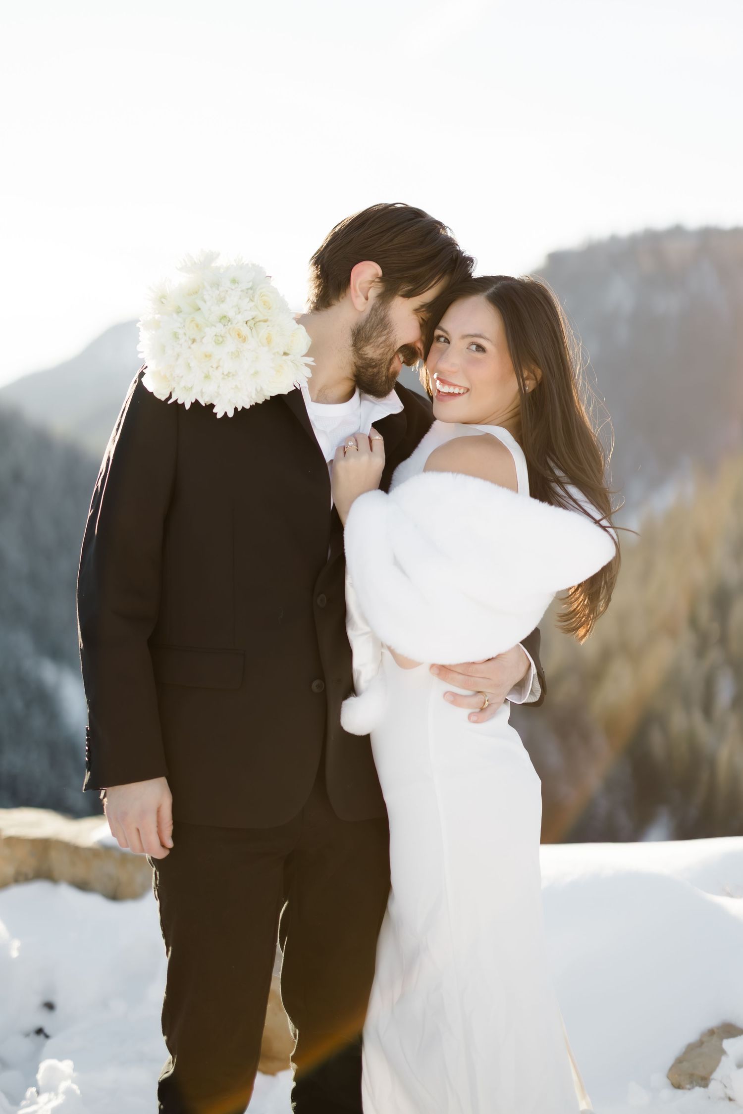 Bride and groom embrace in snowy mountain setting, she wears white fur wrap holding bouquet.