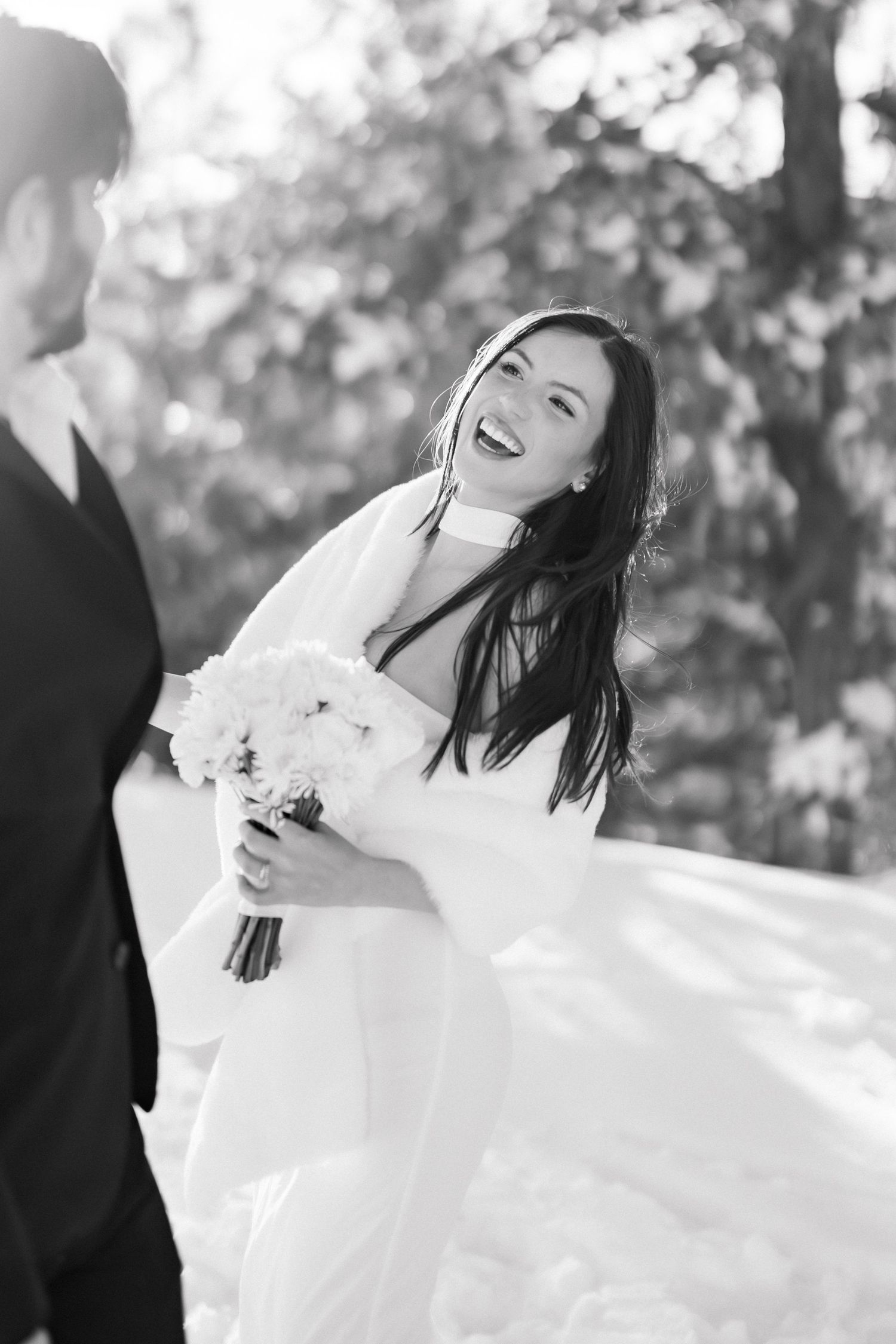 Black and white photo of laughing bride in flowing wedding dress holding bouquet with groom in winter forest.