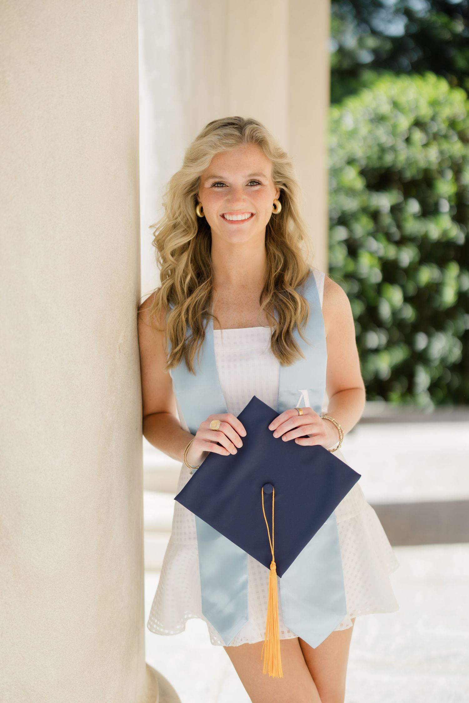 Smiling blonde woman in light blue dress holding navy graduation cap with gold tassel, leaning against white column outdoors.