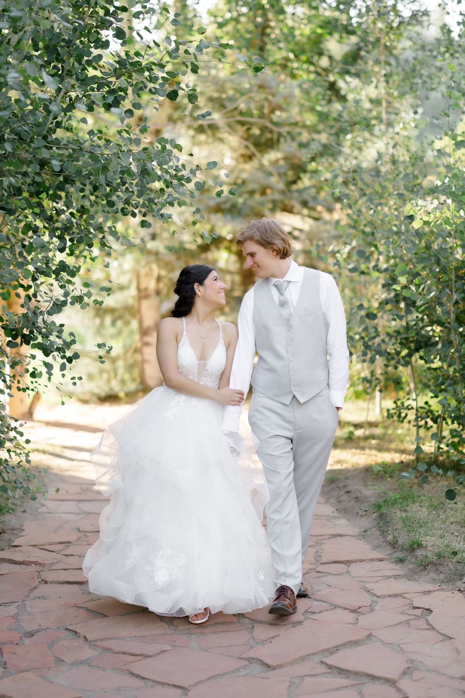 Bride and groom walk hand-in-hand on stone path surrounded by lush green trees on their wedding day.