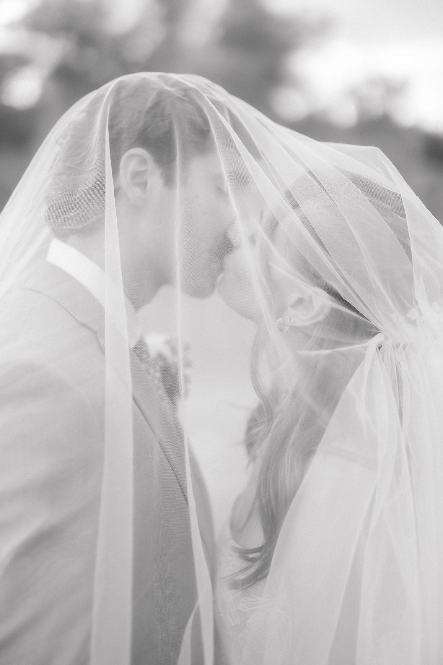 Black and white photo of bride and groom kissing under a flowing wedding veil.