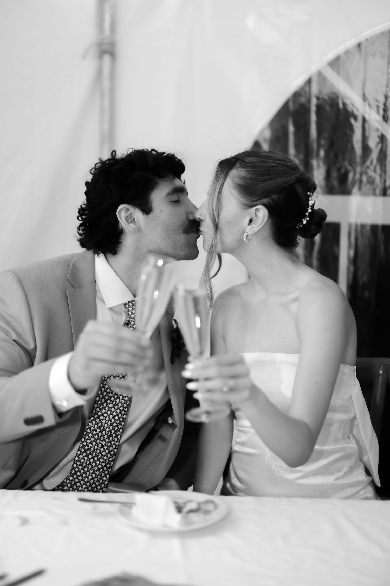 Black and white photo of a bride and groom kissing while clinking champagne glasses at their wedding reception.