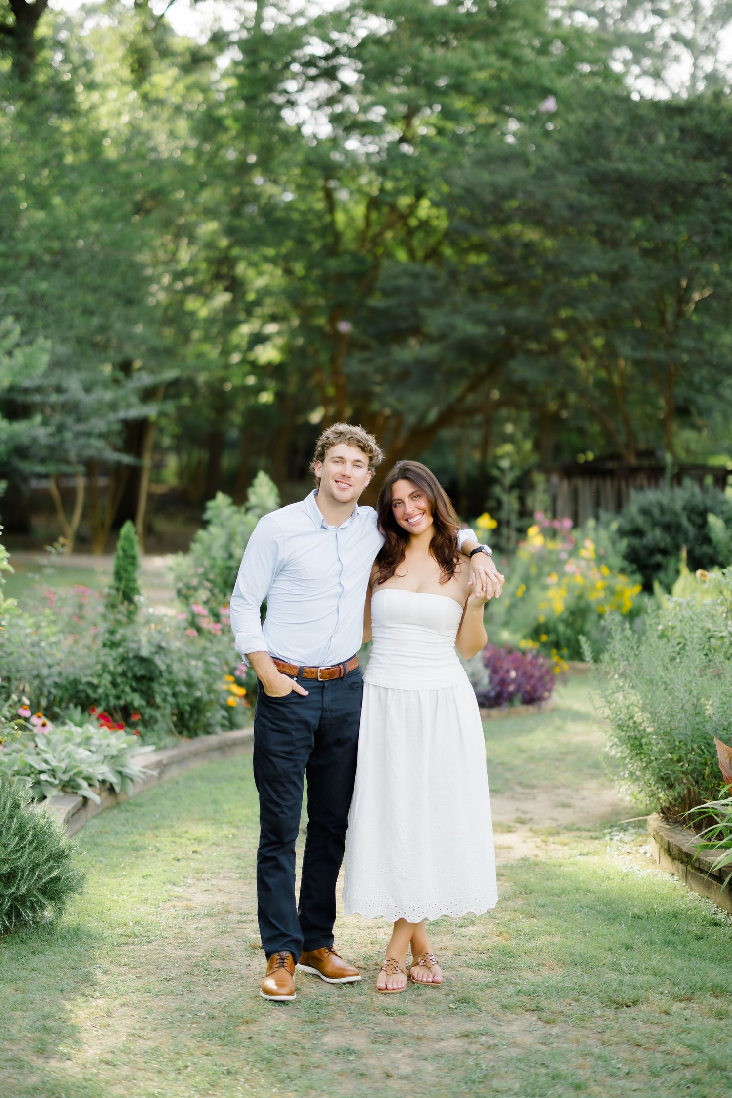 Couple smiling together in a lush garden, man in blue shirt and navy pants, woman in white strapless midi dress.