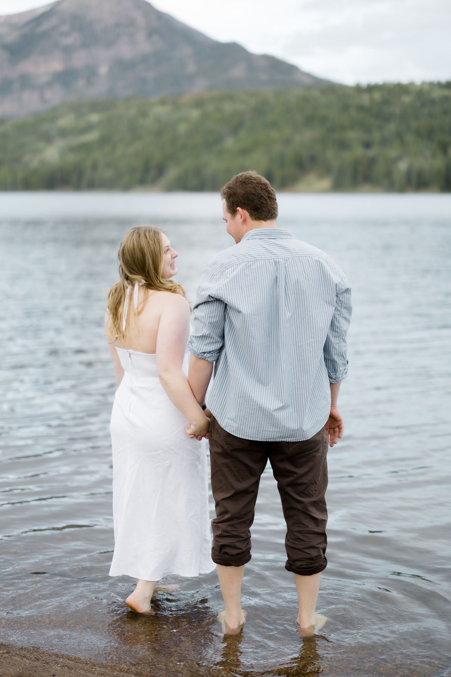 Couple holding hands wading barefoot in a mountain lake, woman in white dress and man in blue shirt and brown pants.