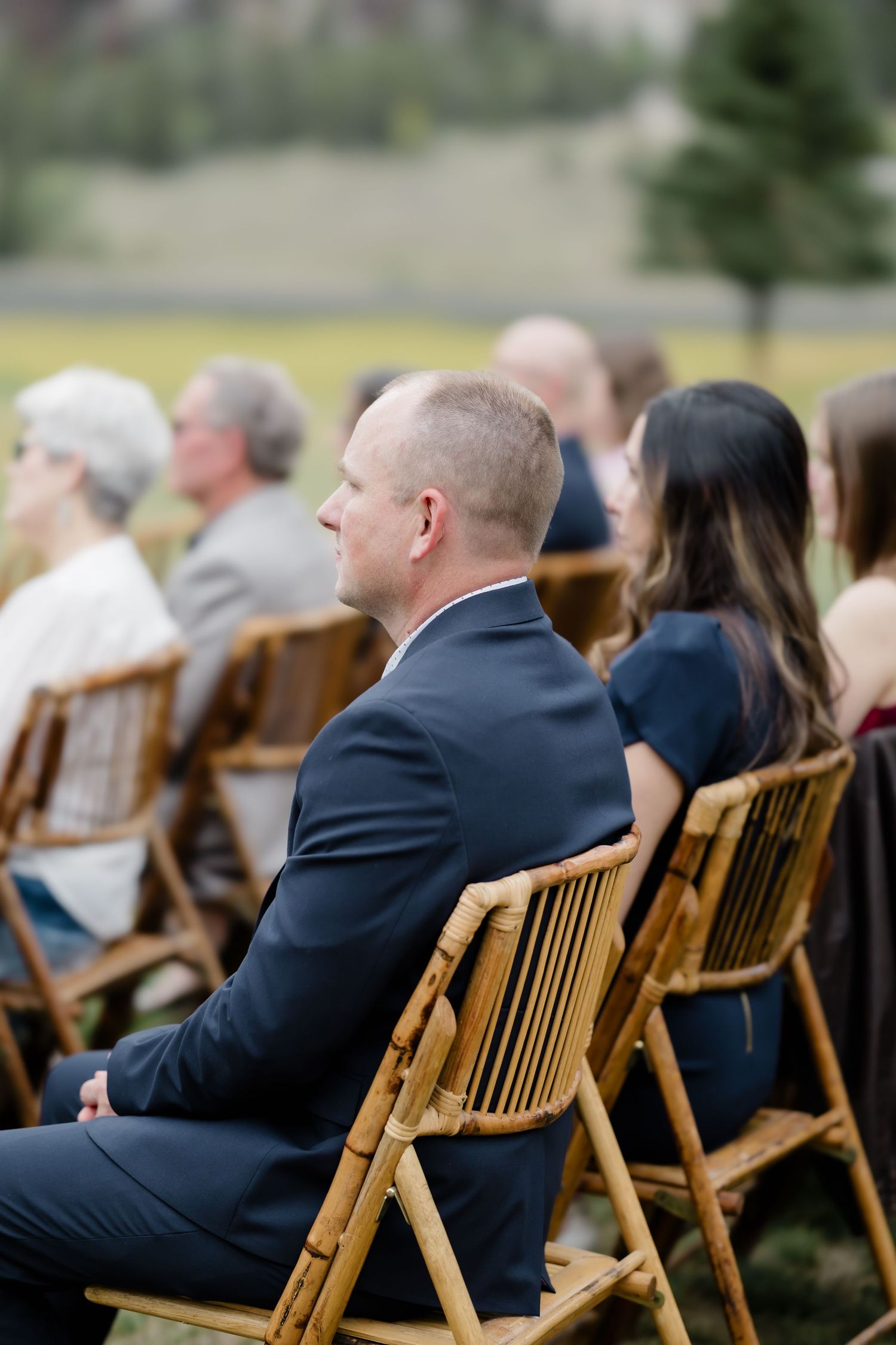 Wedding guests seated in bamboo folding chairs during an outdoor ceremony, man in navy suit in foreground.