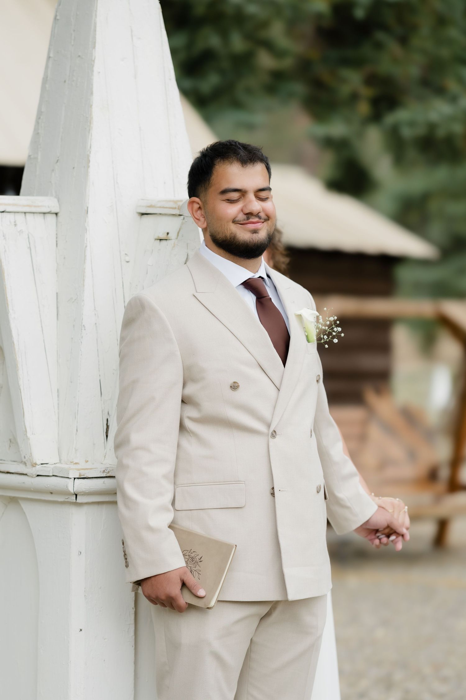 Groom in beige suit with boutonniere holding a book, posing outdoors near white architecture.