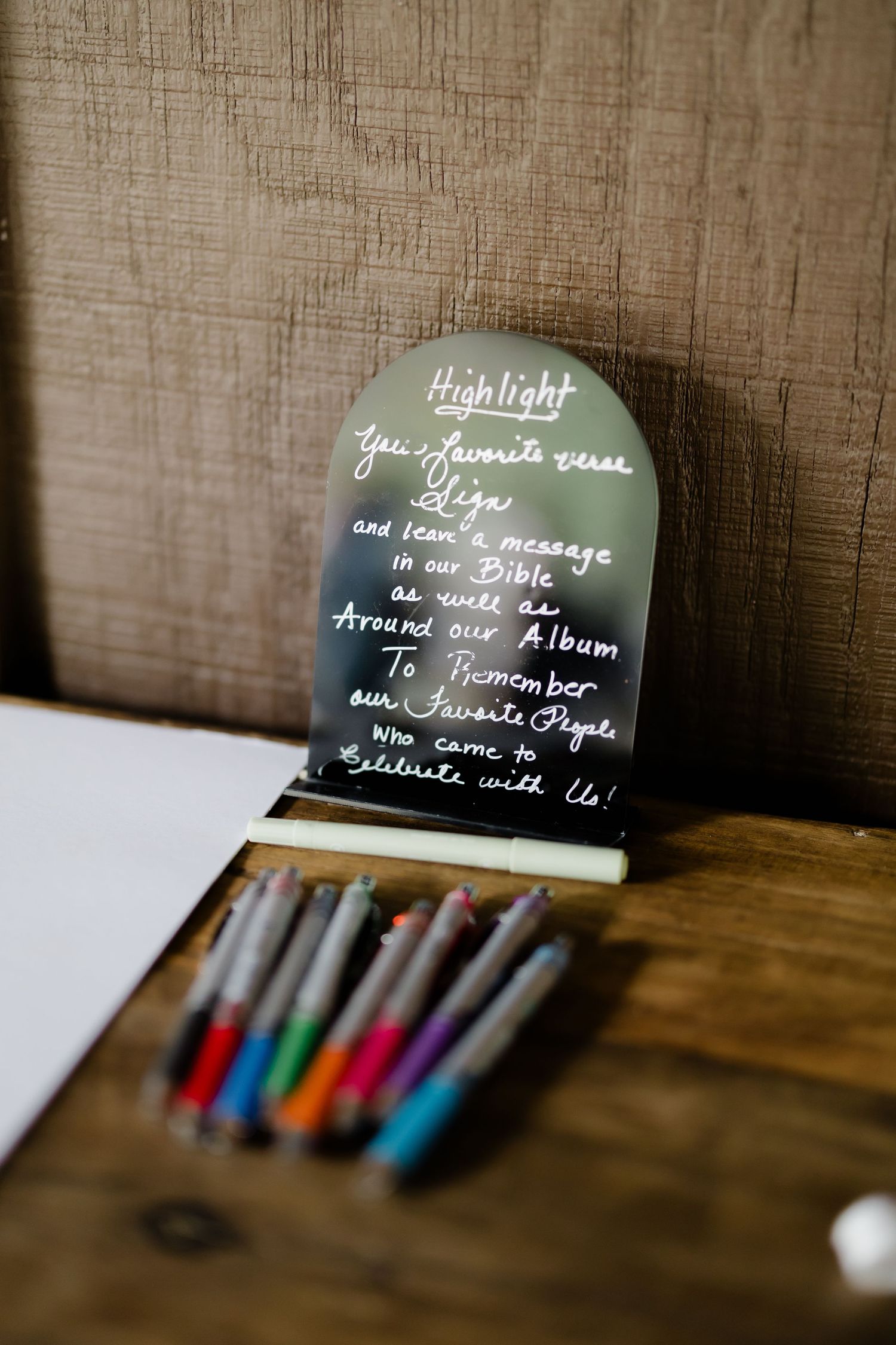 A decorative acrylic sign with handwritten calligraphy inviting wedding guests to highlight a Bible verse, surrounded by colorful pens.