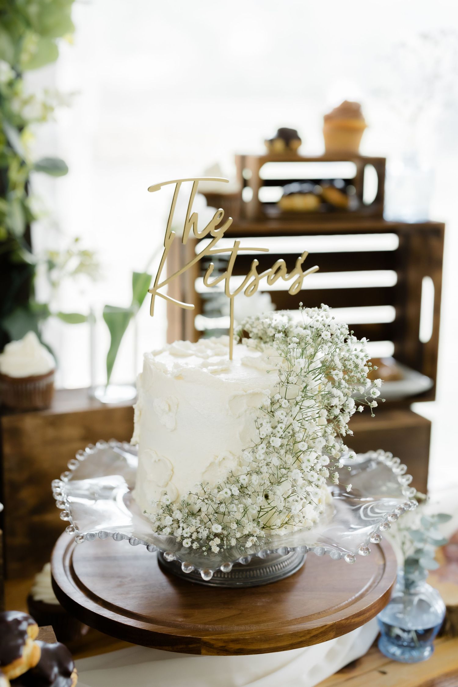 White wedding cake with baby's breath flowers and gold The Kitsas topper on glass stand.
