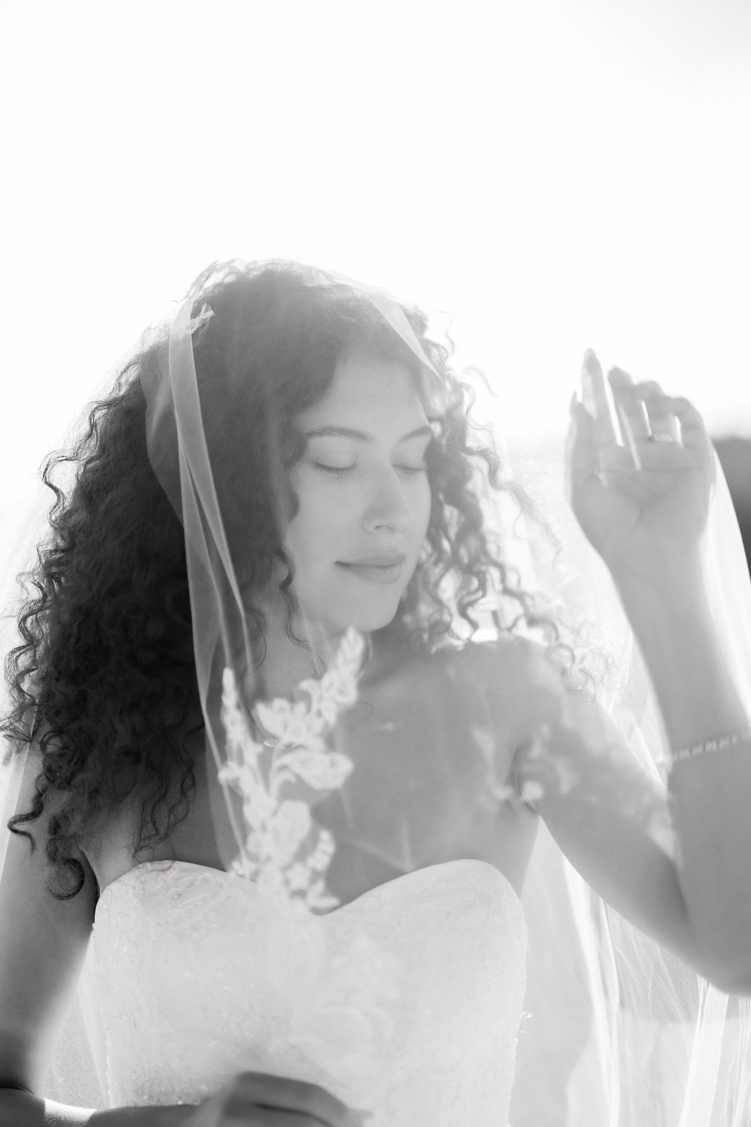 Black and white portrait of a curly-haired bride in a white dress and veil, eyes closed, holding a floral bouquet.