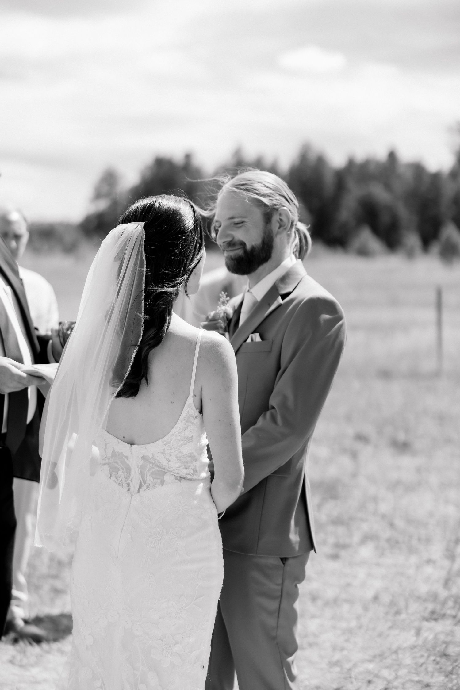 Black and white photo of a smiling groom holding his bride's hands during an outdoor wedding ceremony in a field.