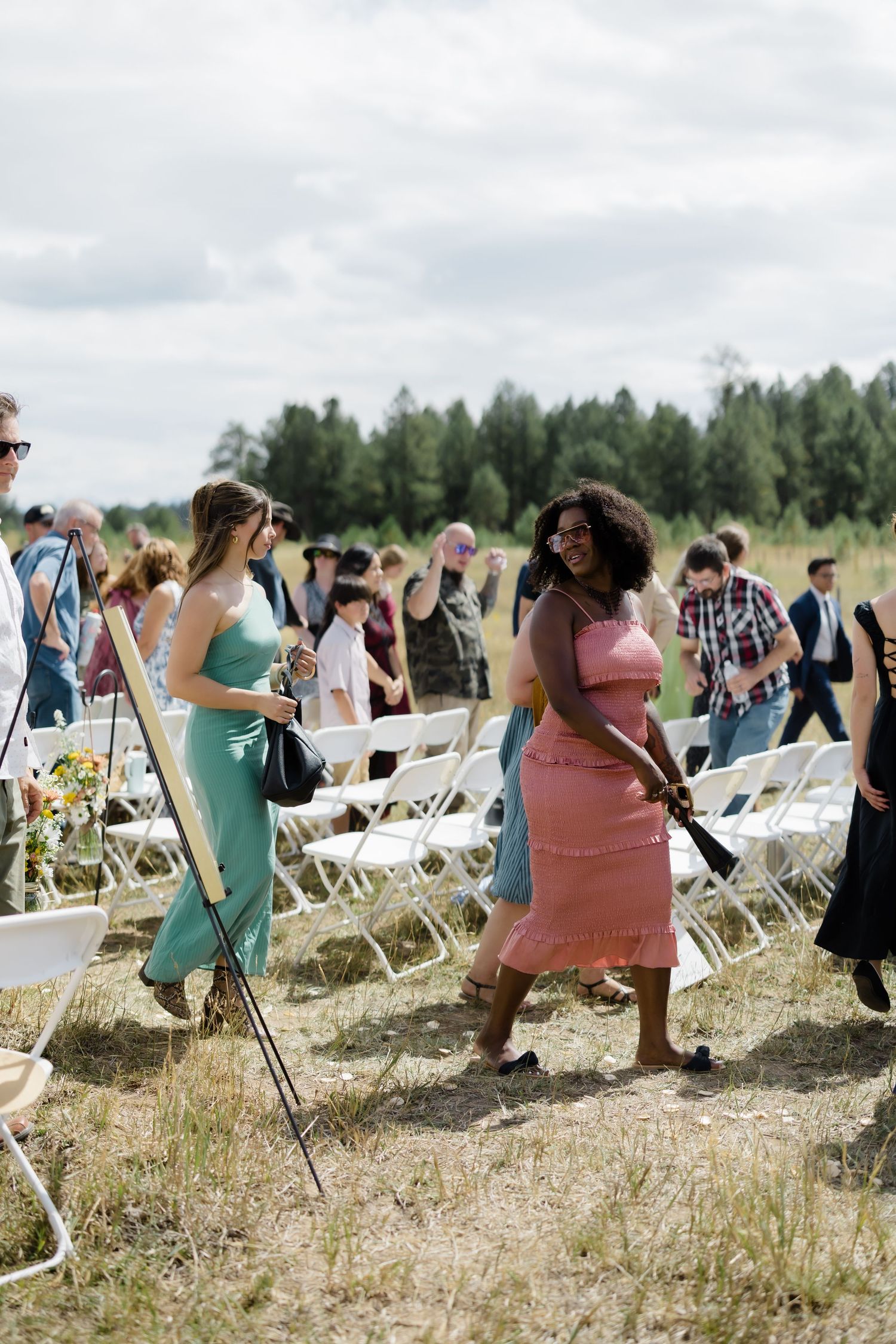 Guests in summer dresses walk among white chairs at an outdoor wedding ceremony in a field.
