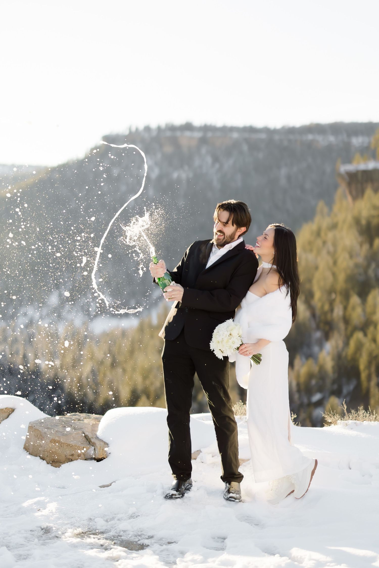 Newlyweds celebrate with champagne spray on snowy mountain overlook, bride in white gown.