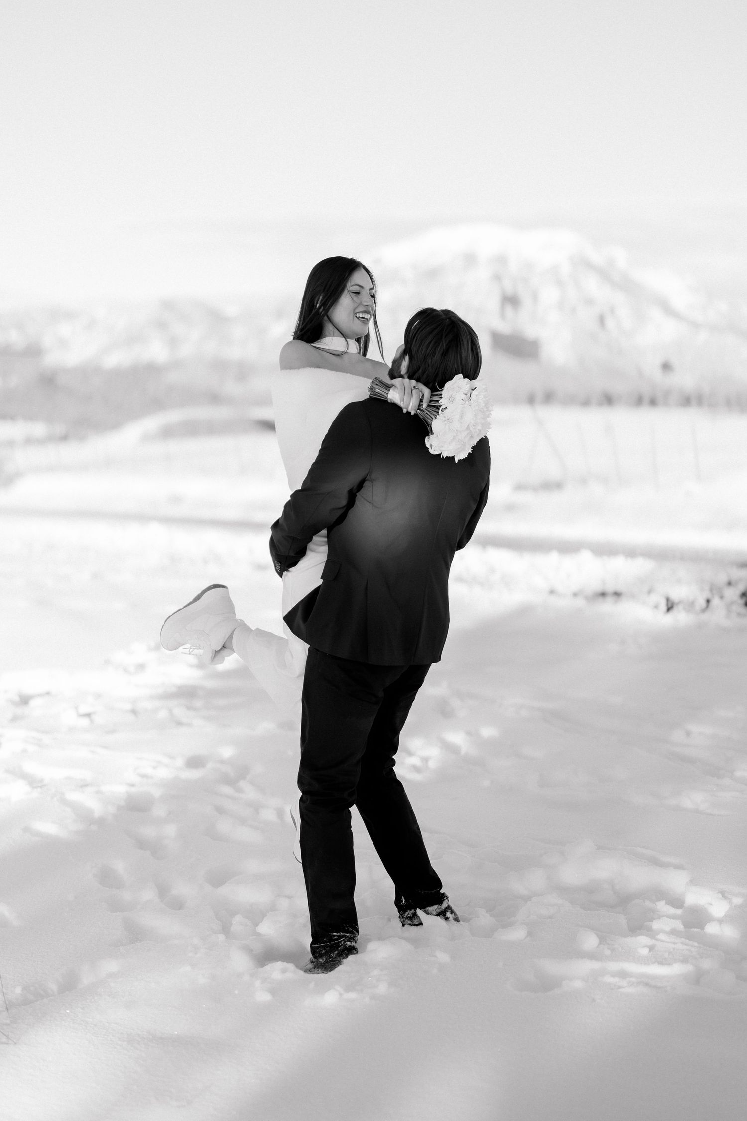 Groom lifts smiling bride in snowy winter landscape with mountain backdrop, romantic black and white wedding photo.
