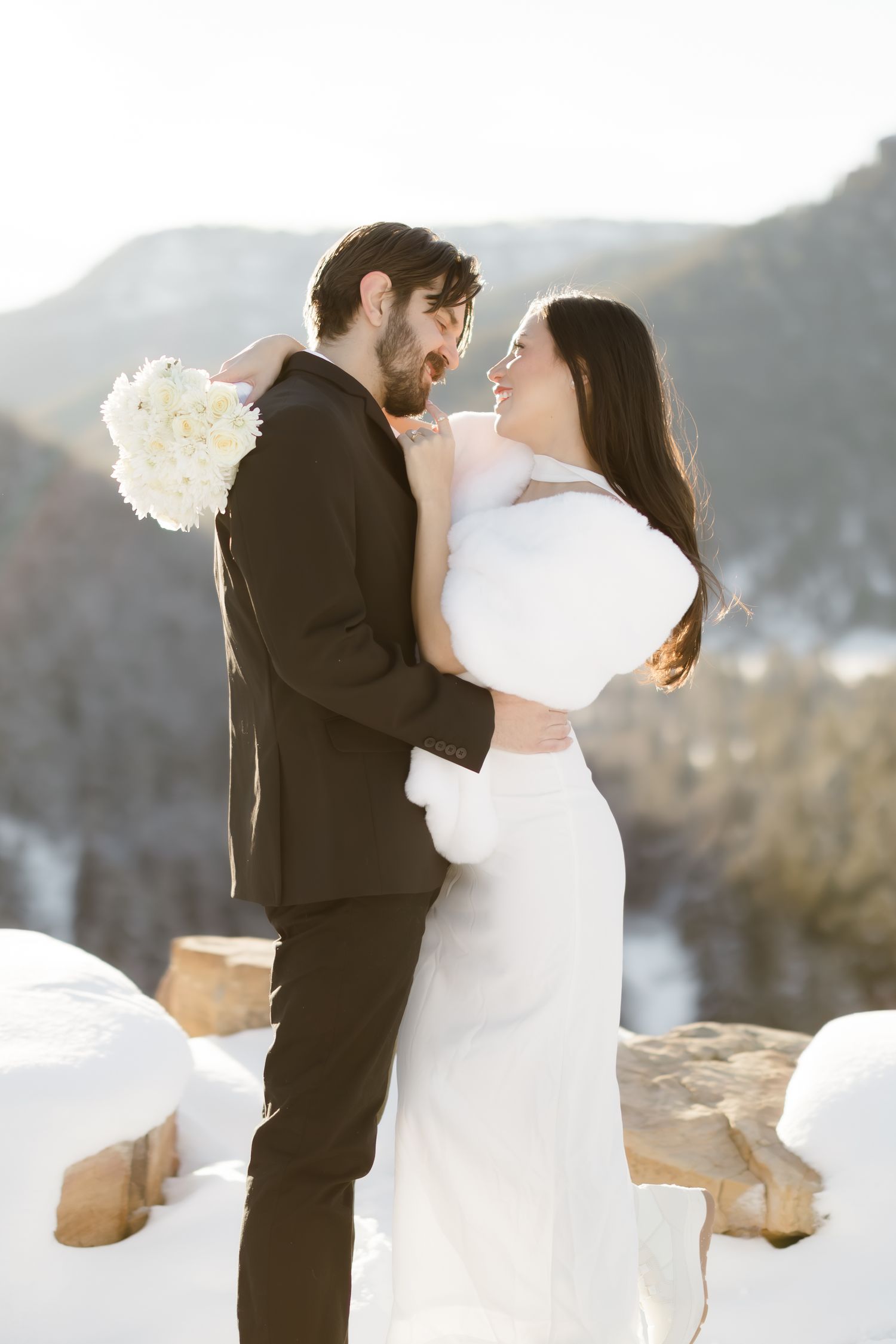 Smiling bride and groom embrace outdoors in snowy mountains, bride in white gown with fur wrap holding bouquet.