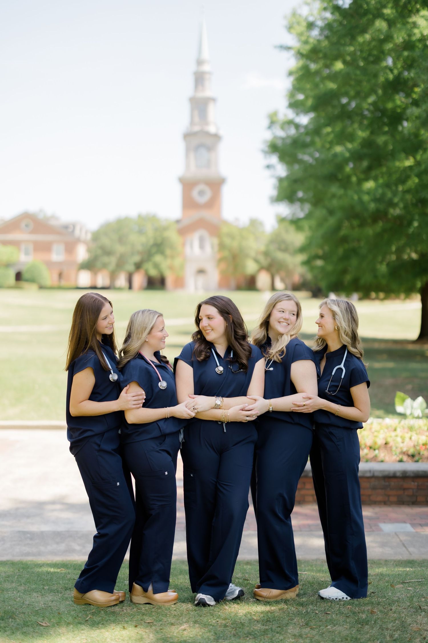 Five female nursing graduates in navy scrubs with stethoscopes pose together on a college campus.