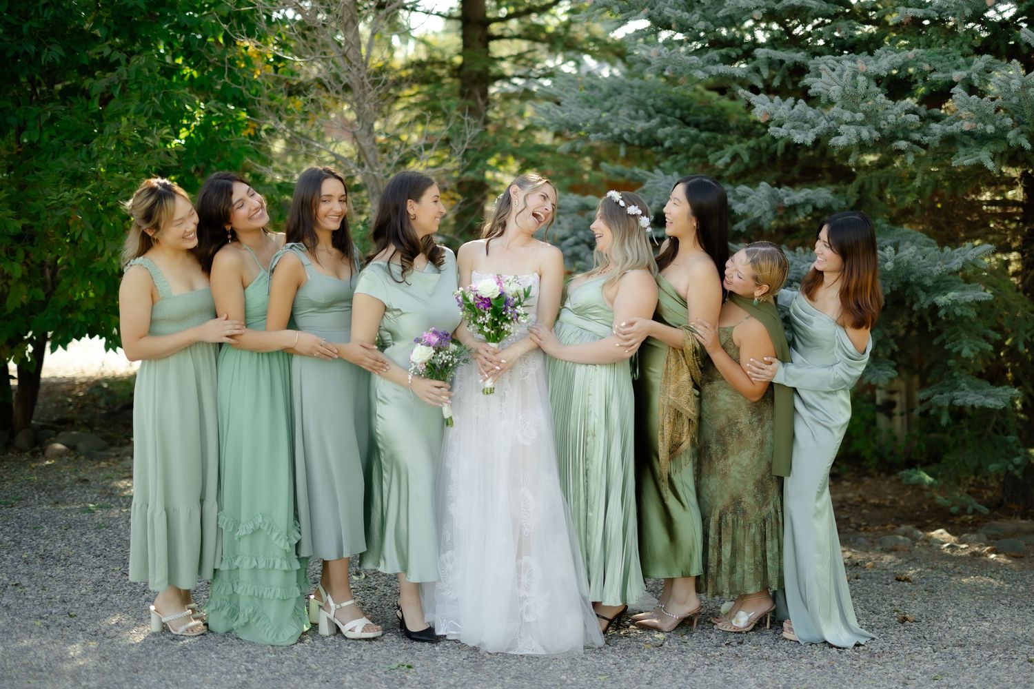 Bride in white lace gown with bridesmaids in sage green dresses laughing together outdoors at a wedding.