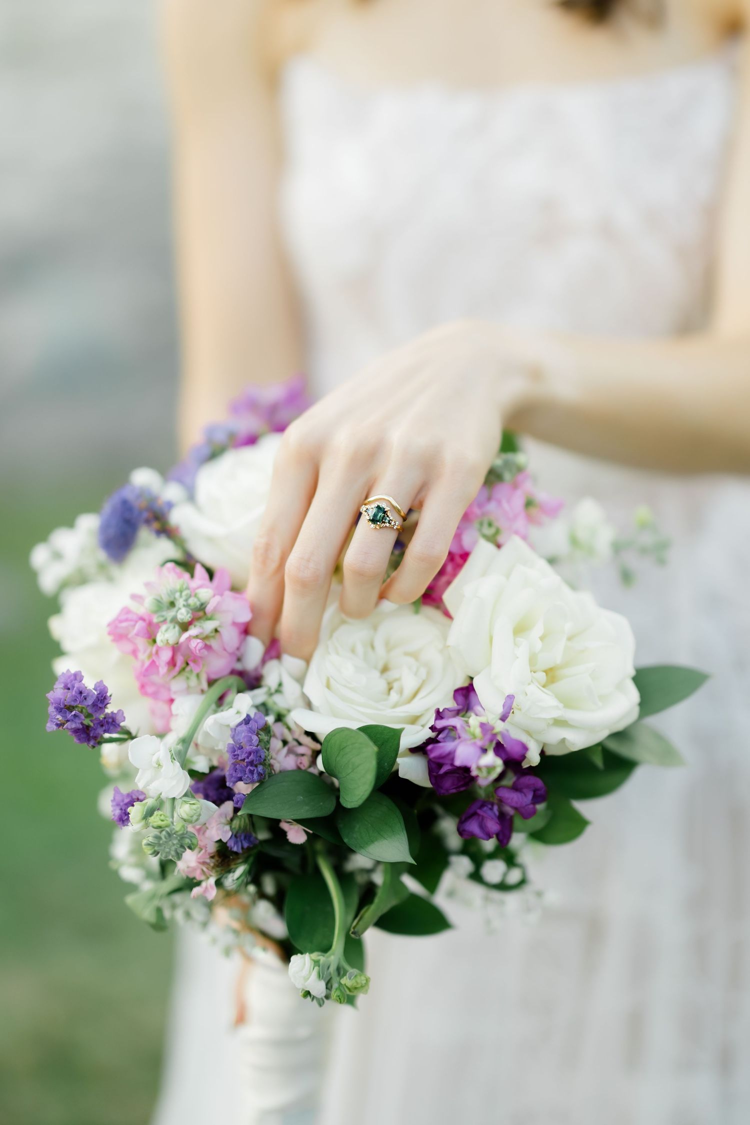 Bride holding a wedding bouquet of white roses and purple flowers, wearing a gold ring with green emerald stone.