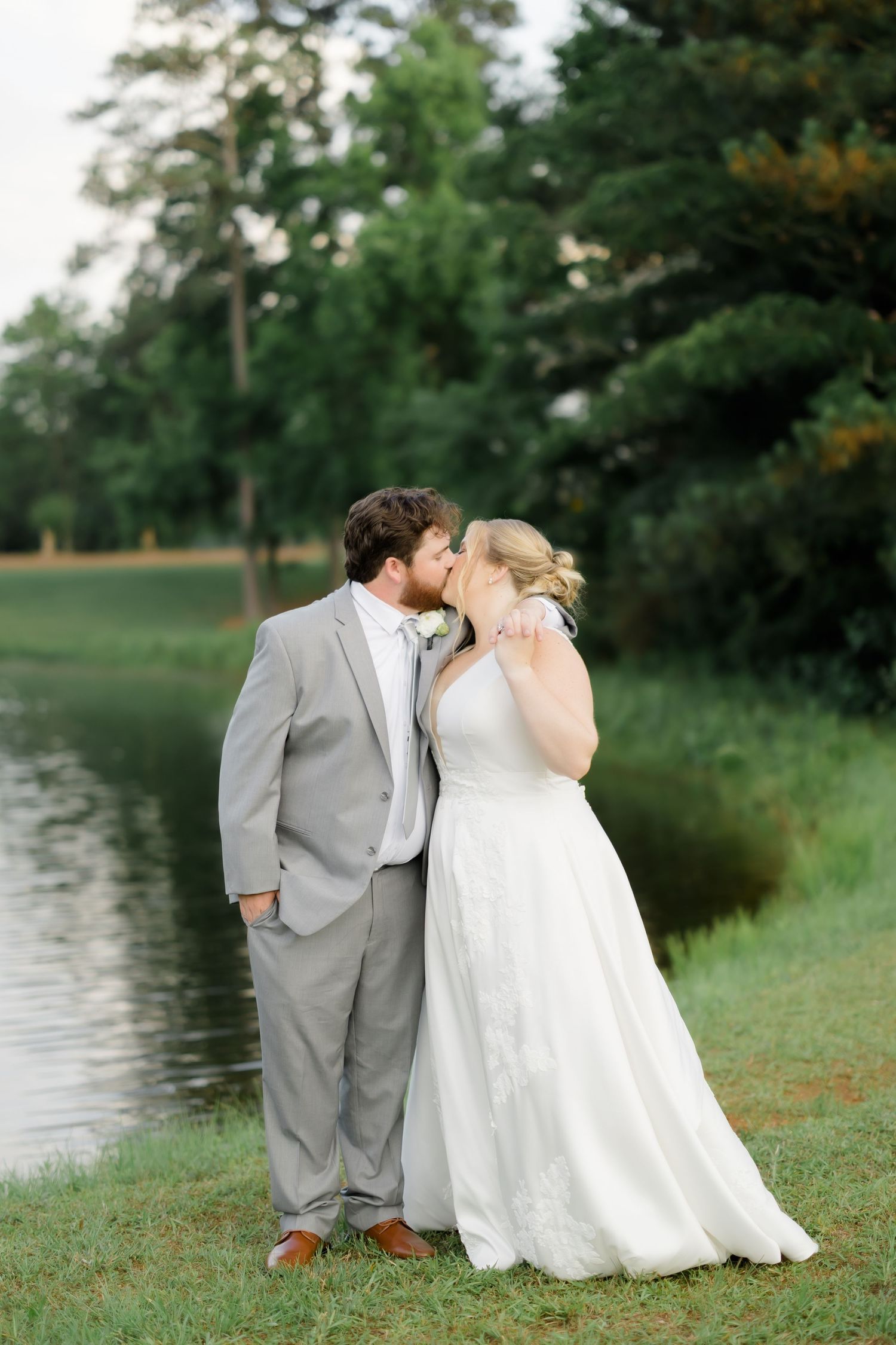 Bride and groom share a romantic kiss by a serene pond, surrounded by lush green trees on their wedding day.