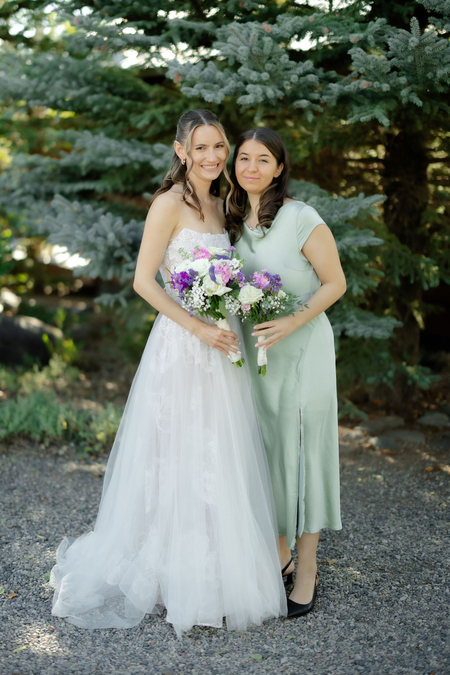 Bride in white gown holds purple bouquet beside bridesmaid in sage green dress, posing outdoors near evergreen trees.