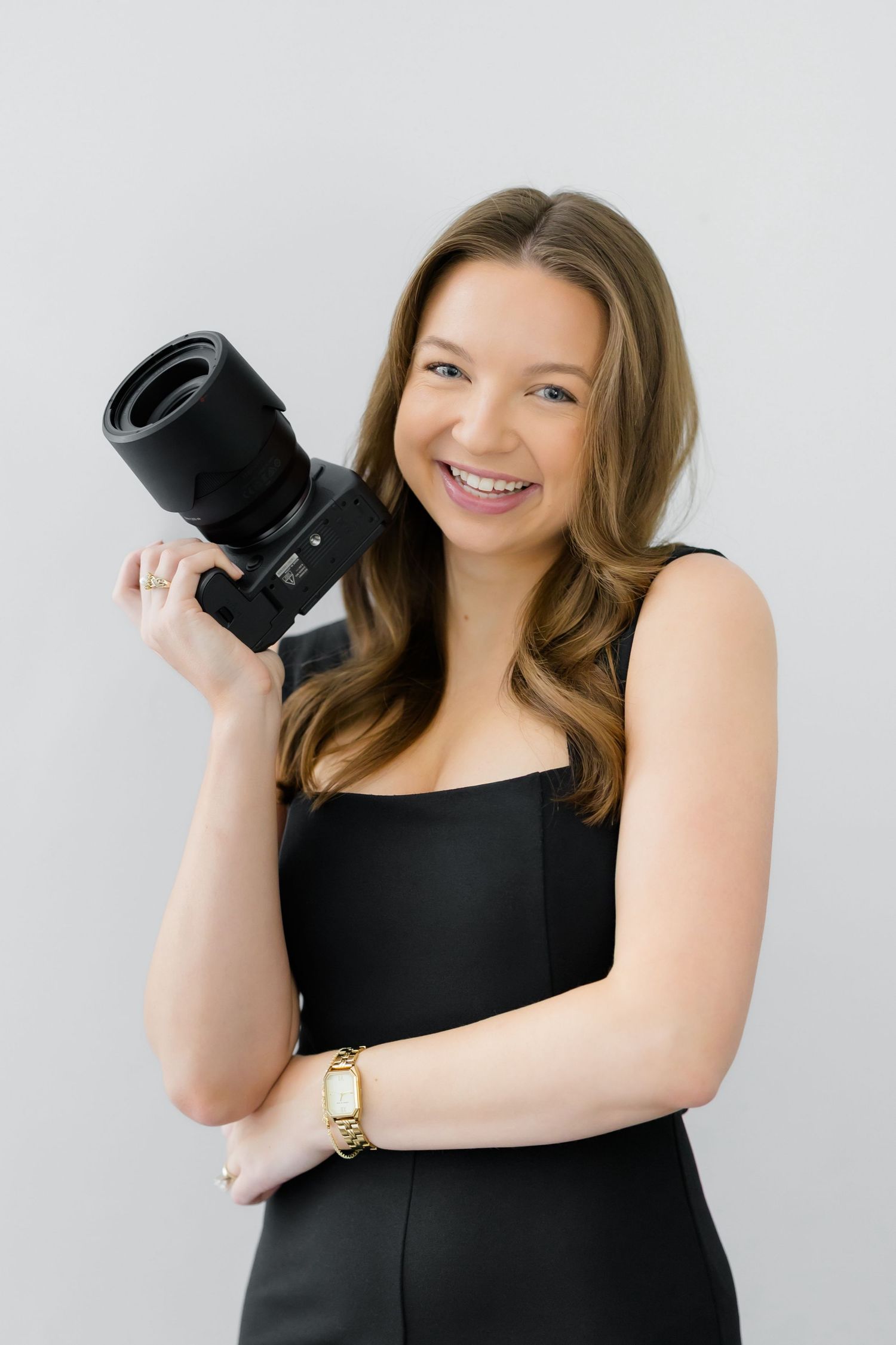 Smiling young woman in black dress holding a professional camera, posing against a light gray background.