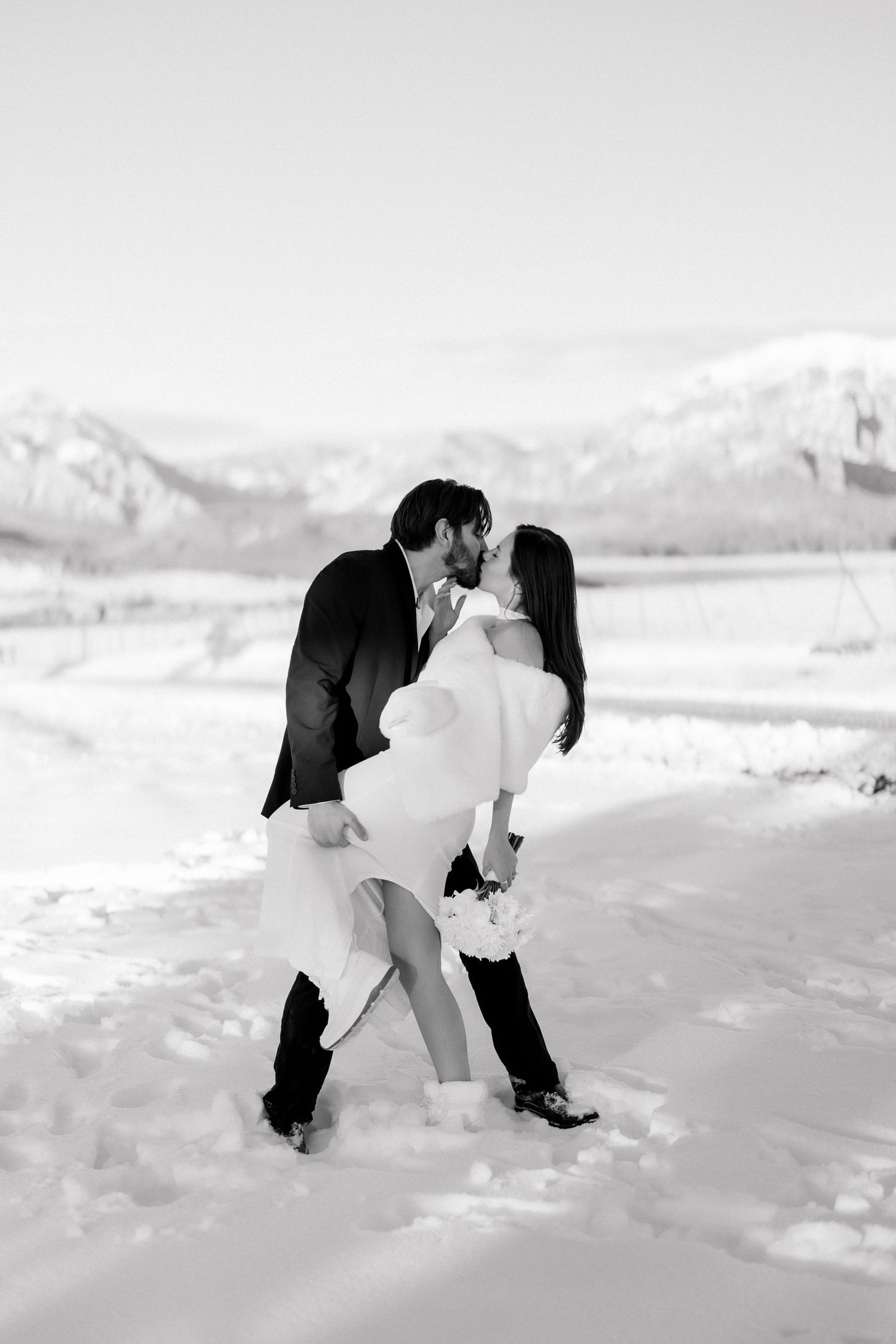 Black and white photo of bride and groom sharing a kiss while dipping in snowy mountain setting.