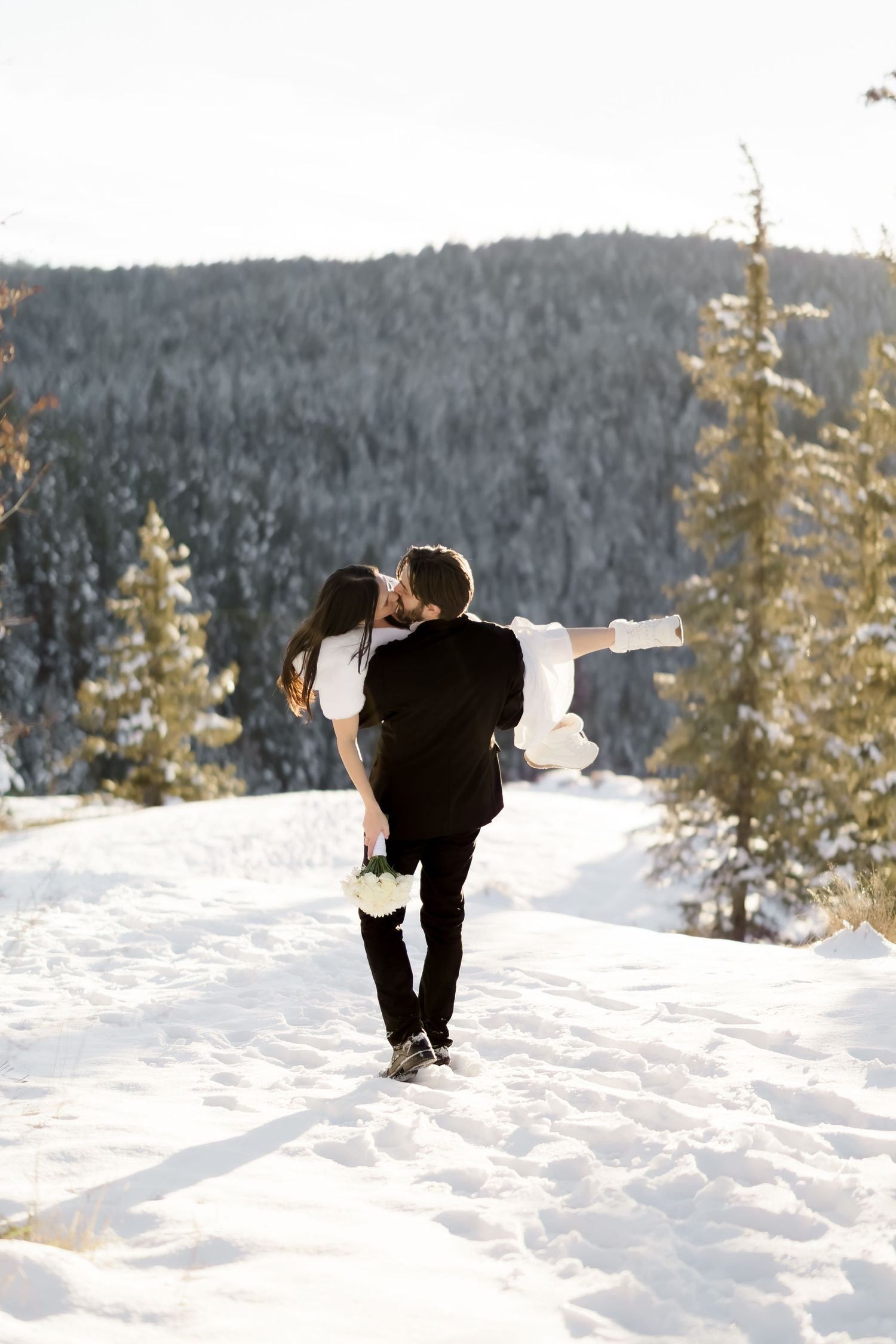 Groom carrying bride in white dress through snowy mountain landscape while sharing a romantic kiss.