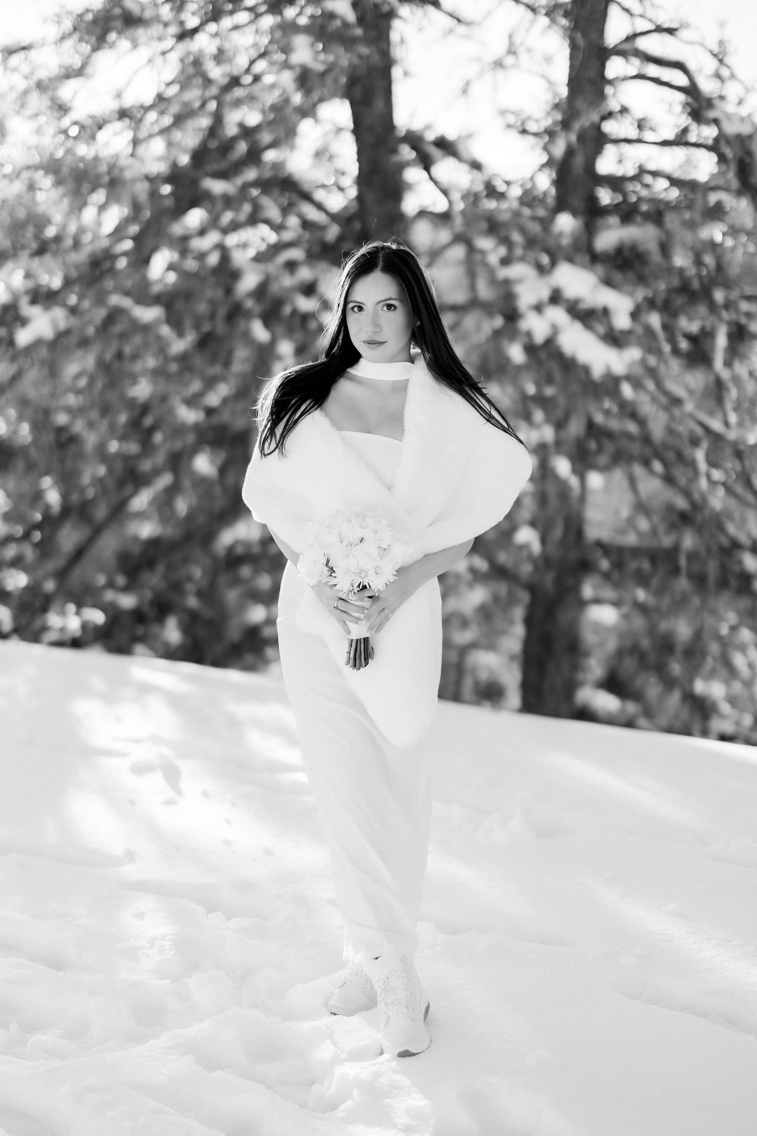 Bride in white gown holds bouquet in snowy winter forest, black and white wedding photo.