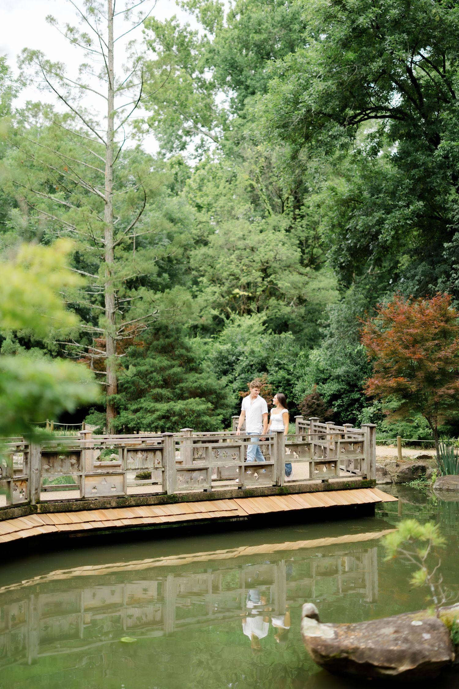 A couple stands together on a wooden bridge over a serene pond surrounded by lush green trees.