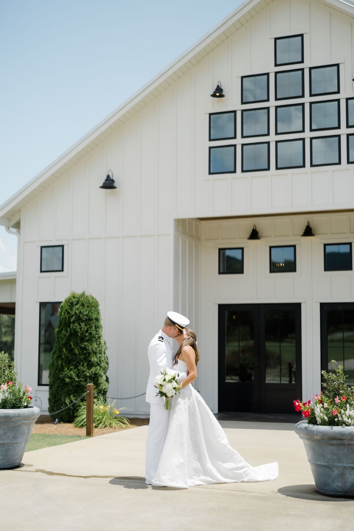 A bride and groom share a kiss outside a white barn wedding venue, with the groom in a naval uniform and bride in a gown.