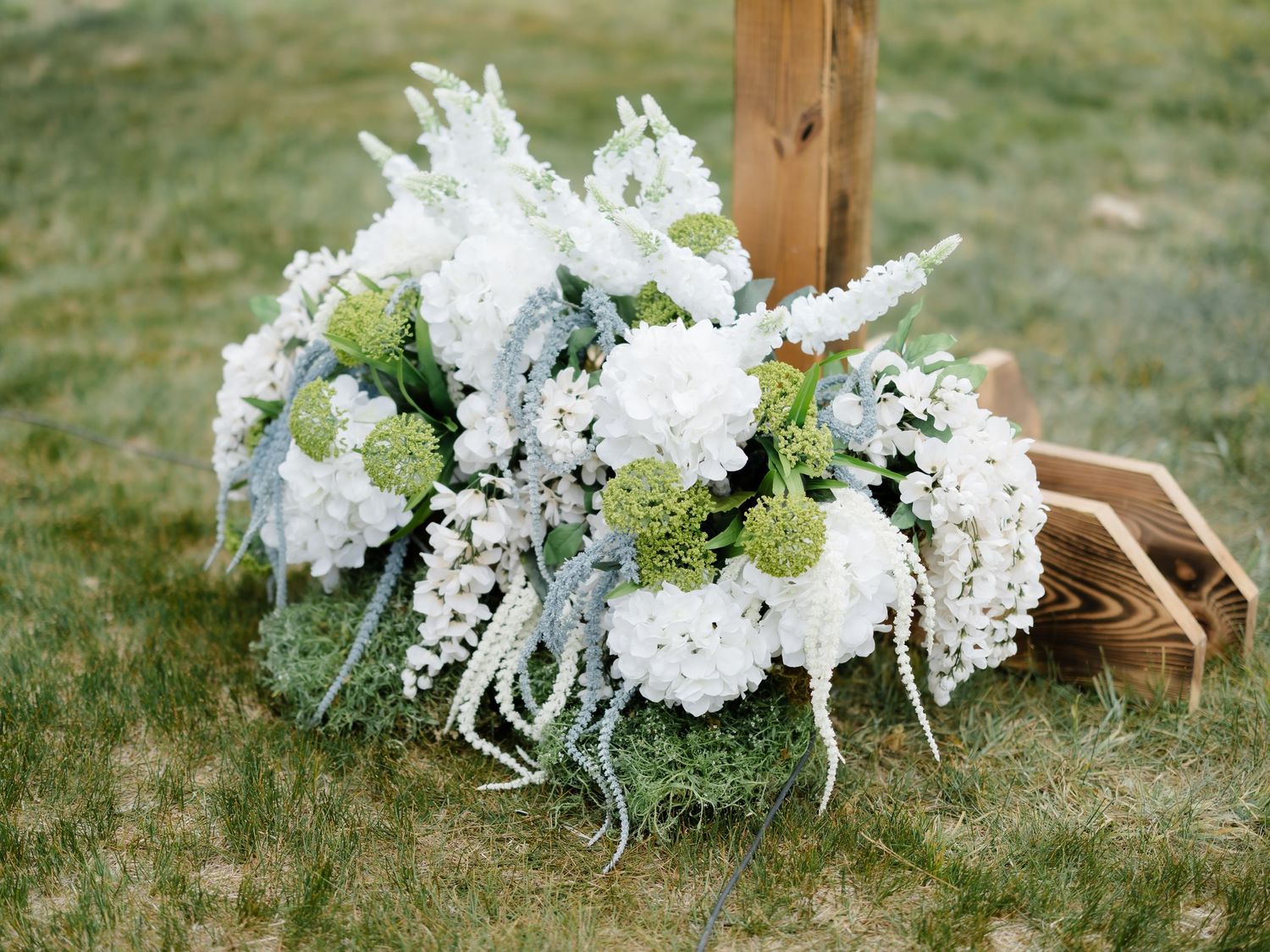 White hydrangea and green floral wedding bouquet arrangement on grass beside a wooden post.