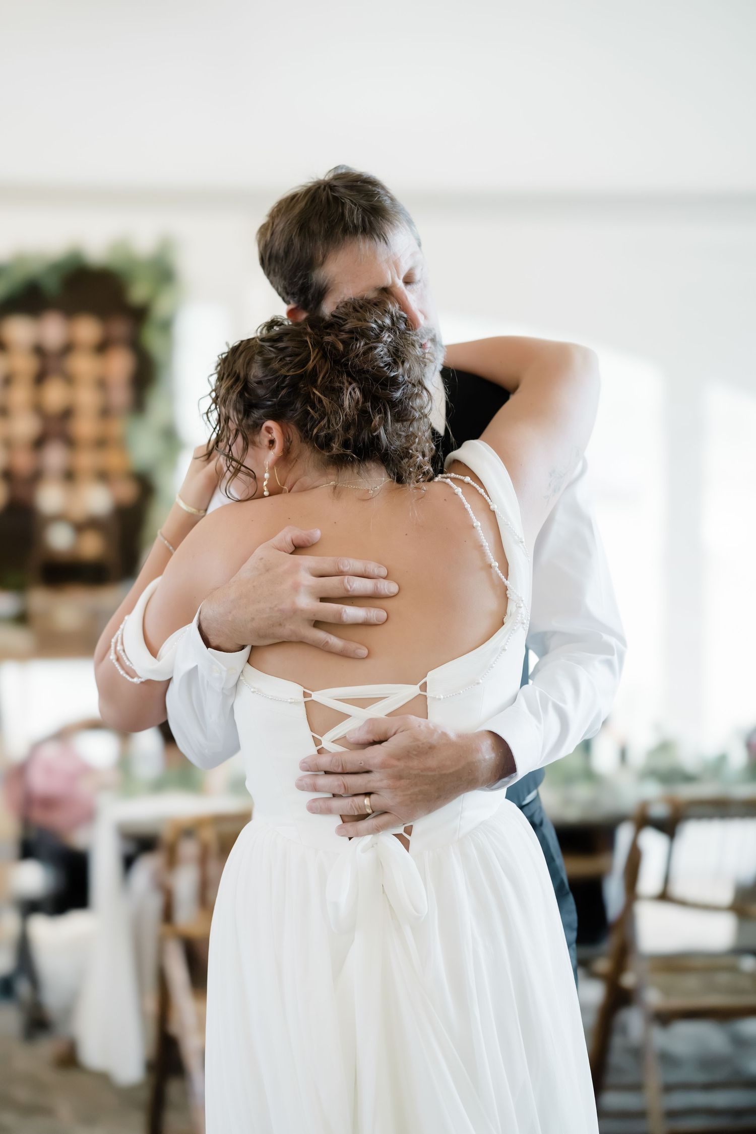 Bride in white off-shoulder gown sharing emotional hug with father during wedding reception dance.