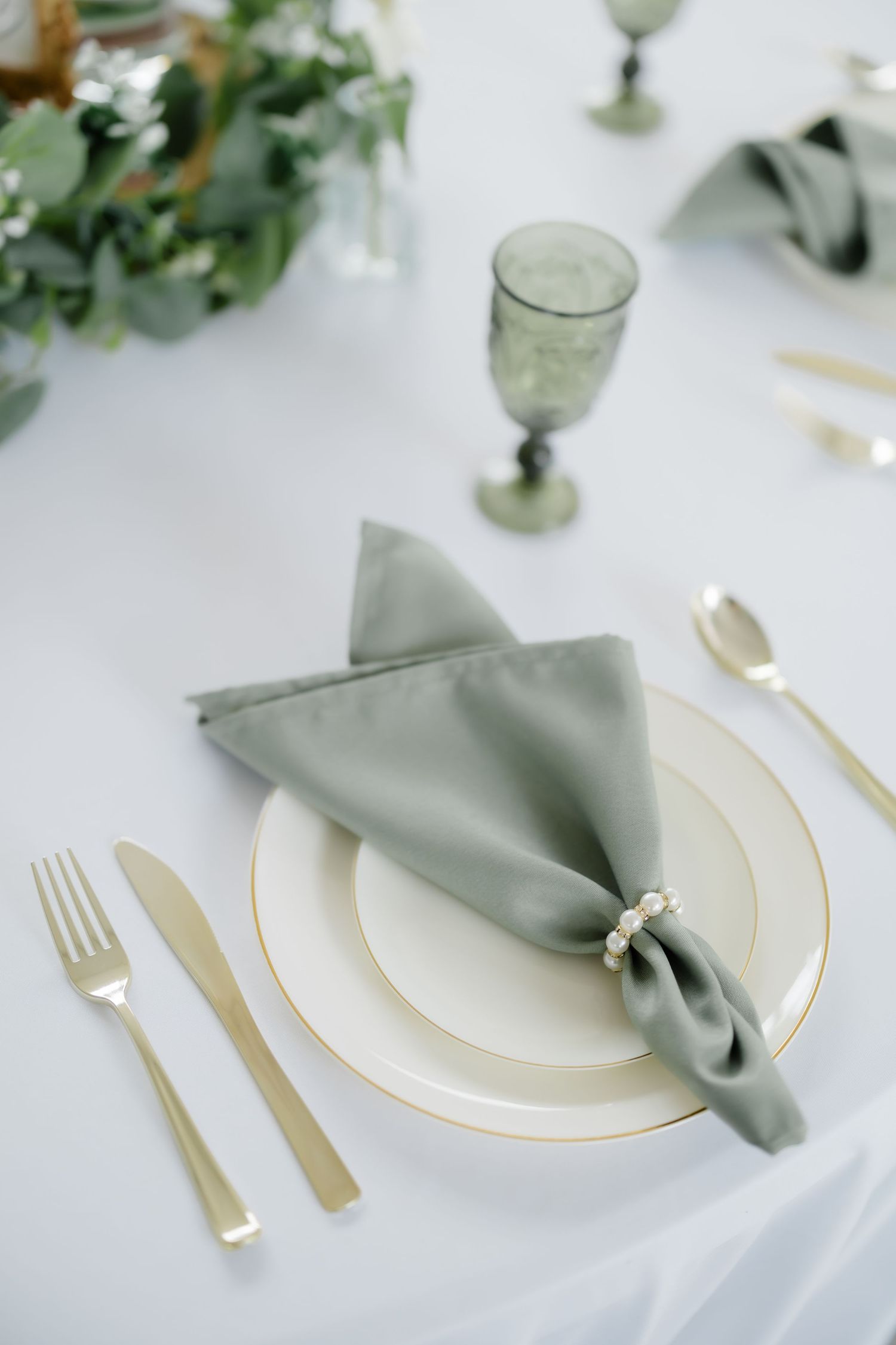 Elegant wedding table setting with gold cutlery, white plates, sage green napkin with pearl ring, and green goblet.