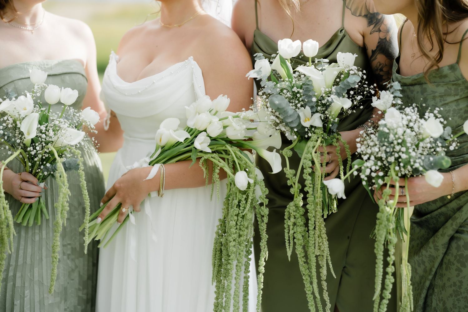 Bride and bridesmaids in sage green dresses holding white tulip and baby's breath bouquets with cascading greenery.