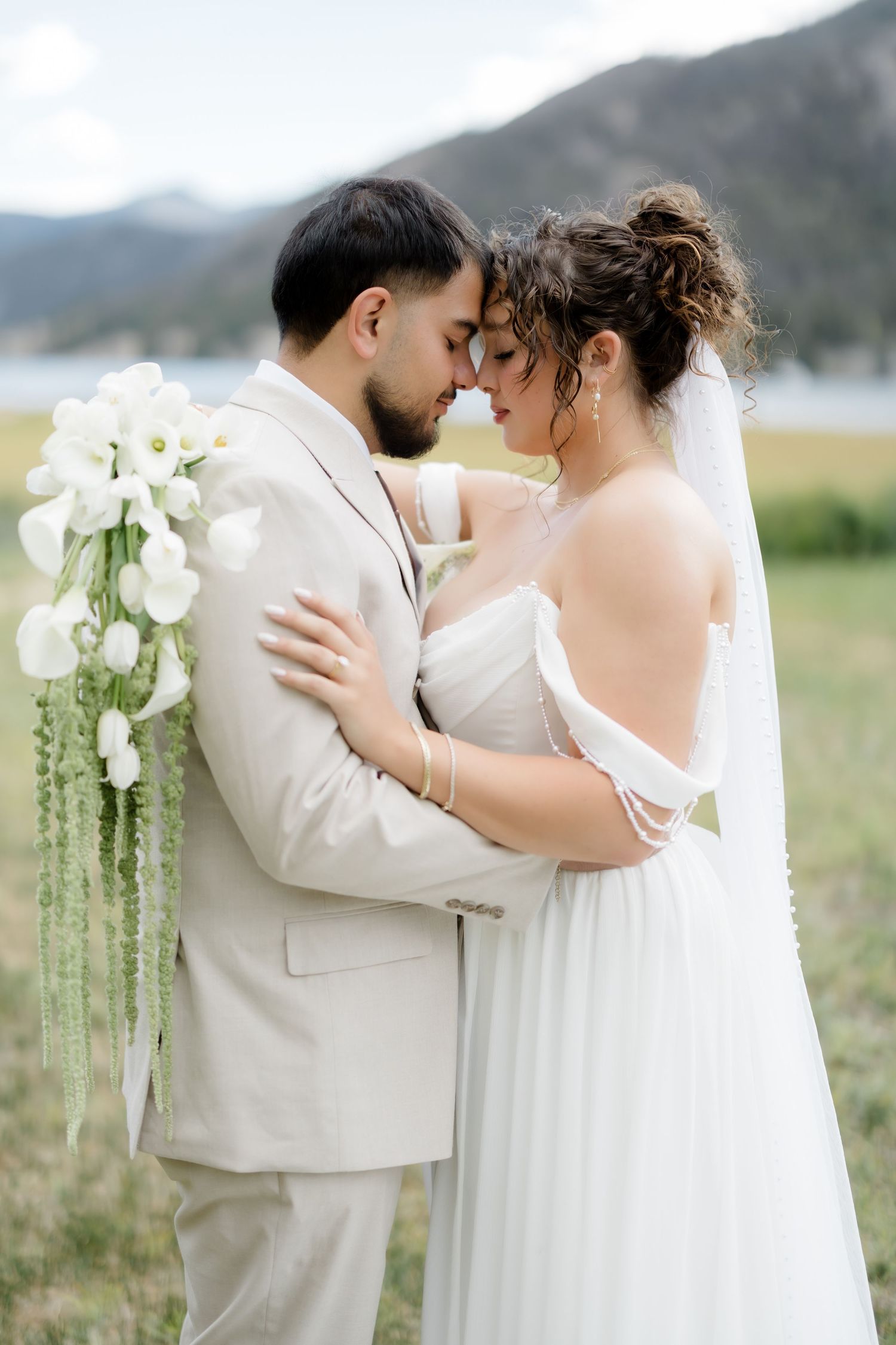Bride and groom embrace outdoors with mountain lake backdrop, bride holding white calla lily bouquet in wedding attire.