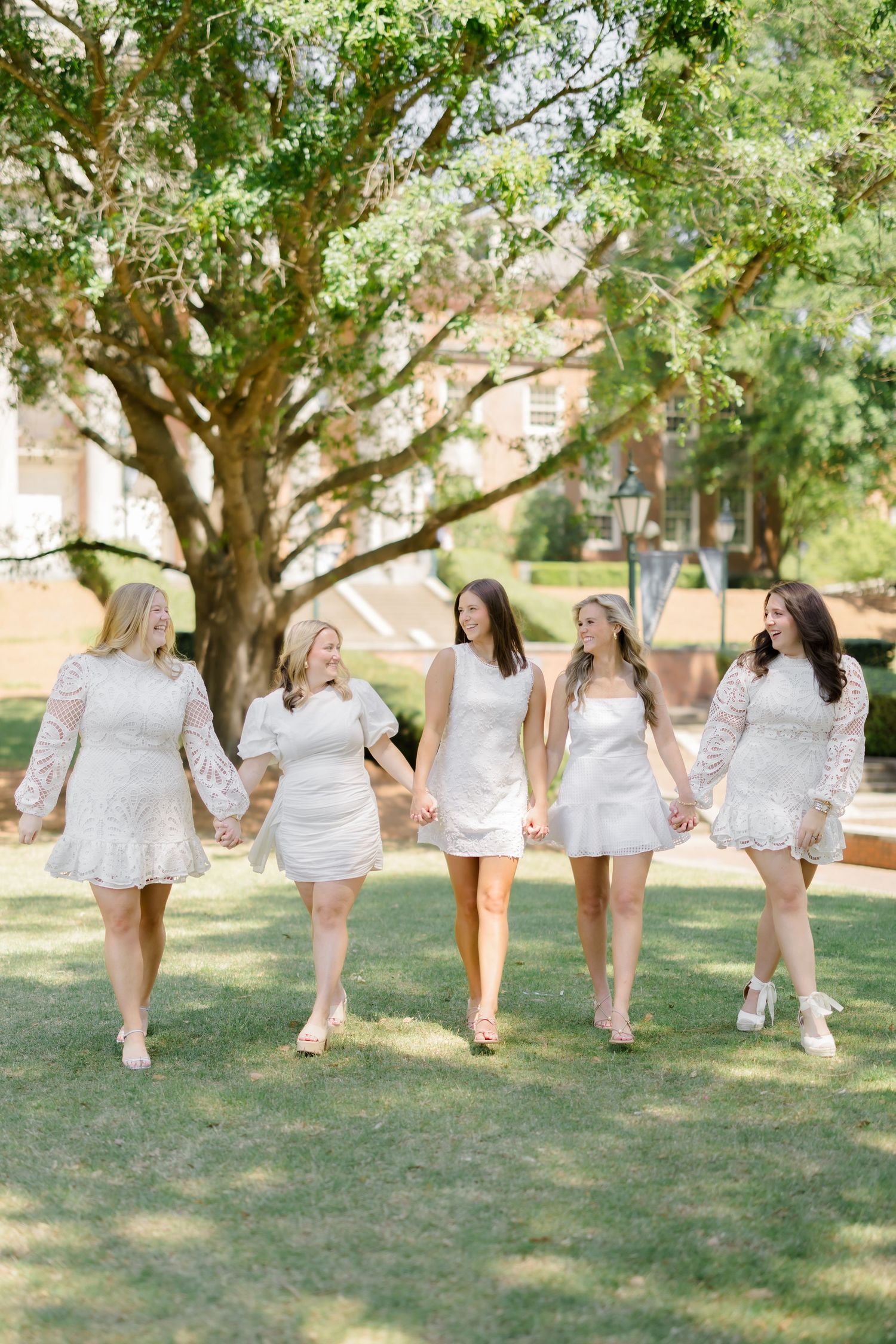 Five women in white dresses walk hand-in-hand on green grass beneath a large tree outdoors.