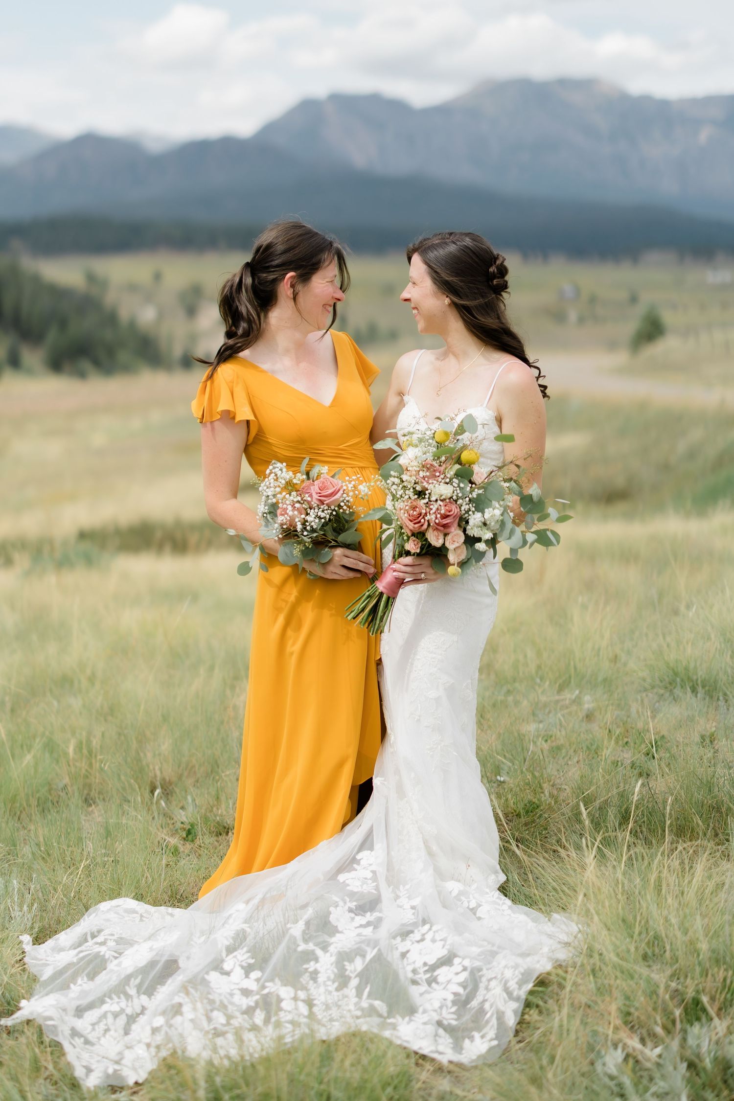 Bride in white lace gown and bridesmaid in yellow dress hold bouquets in a mountain meadow.