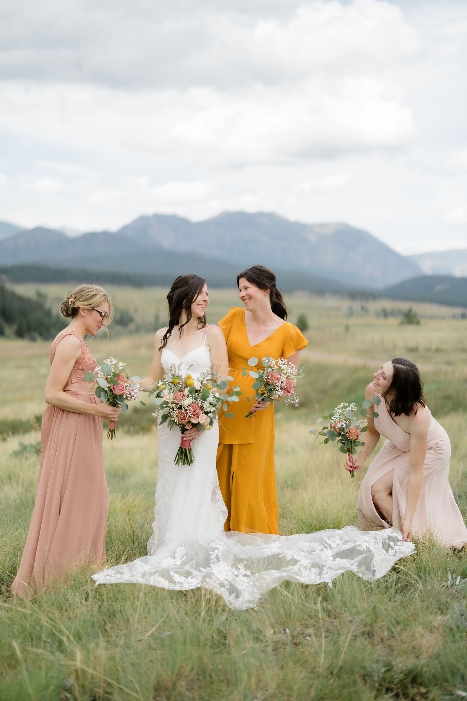 Bride in white gown with three bridesmaids in blush and mustard dresses holding bouquets in a mountain meadow.