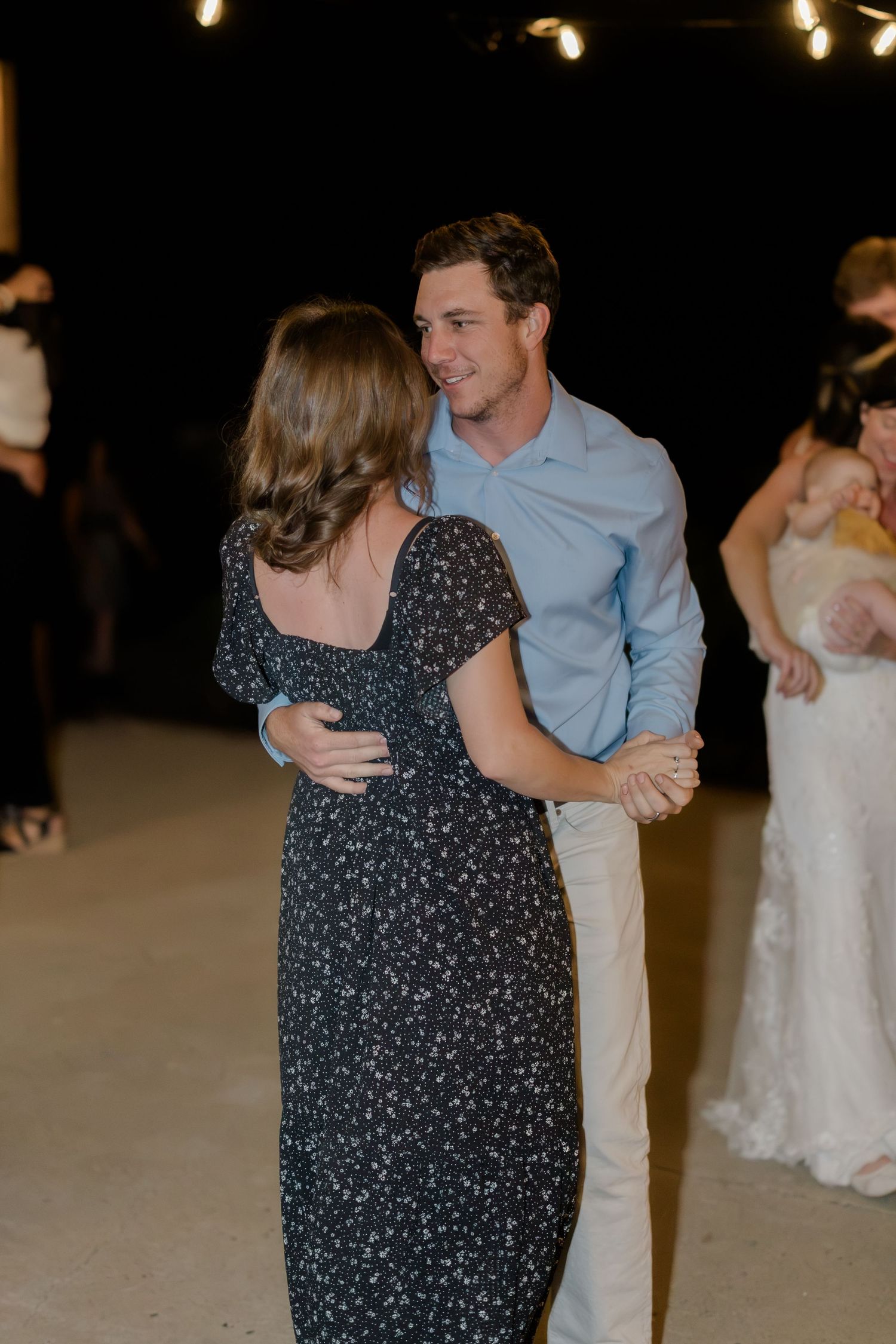 Couple dancing together at a wedding reception, man in blue shirt and woman in floral dress, string lights in background.