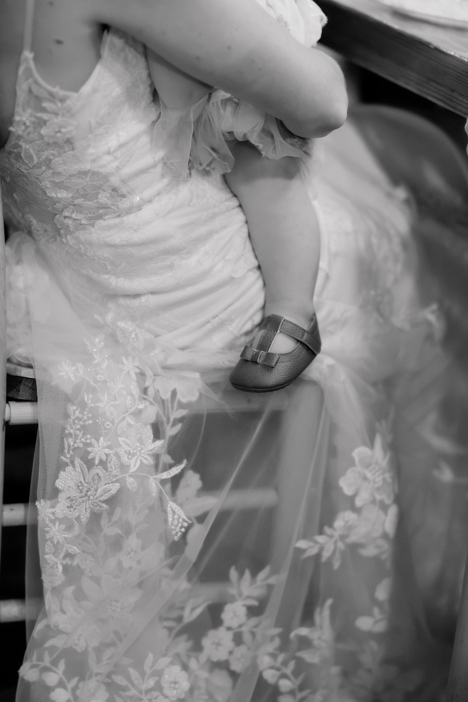 Black and white photo of a bride in lace wedding gown holding a baby with small shoes visible.