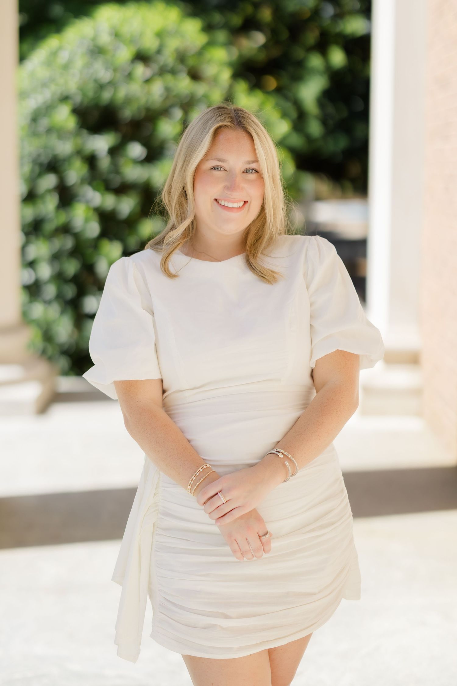 Smiling blonde woman in a white ruched mini dress with puff sleeves, posing outdoors near greenery.
