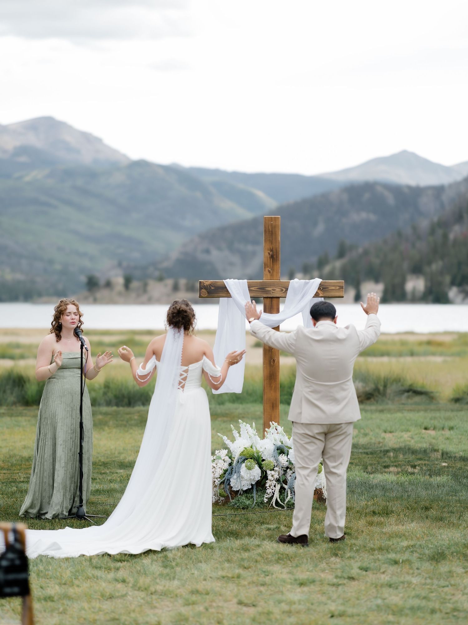 Couple exchange vows at rustic wooden cross altar with mountain lake backdrop at outdoor wedding.