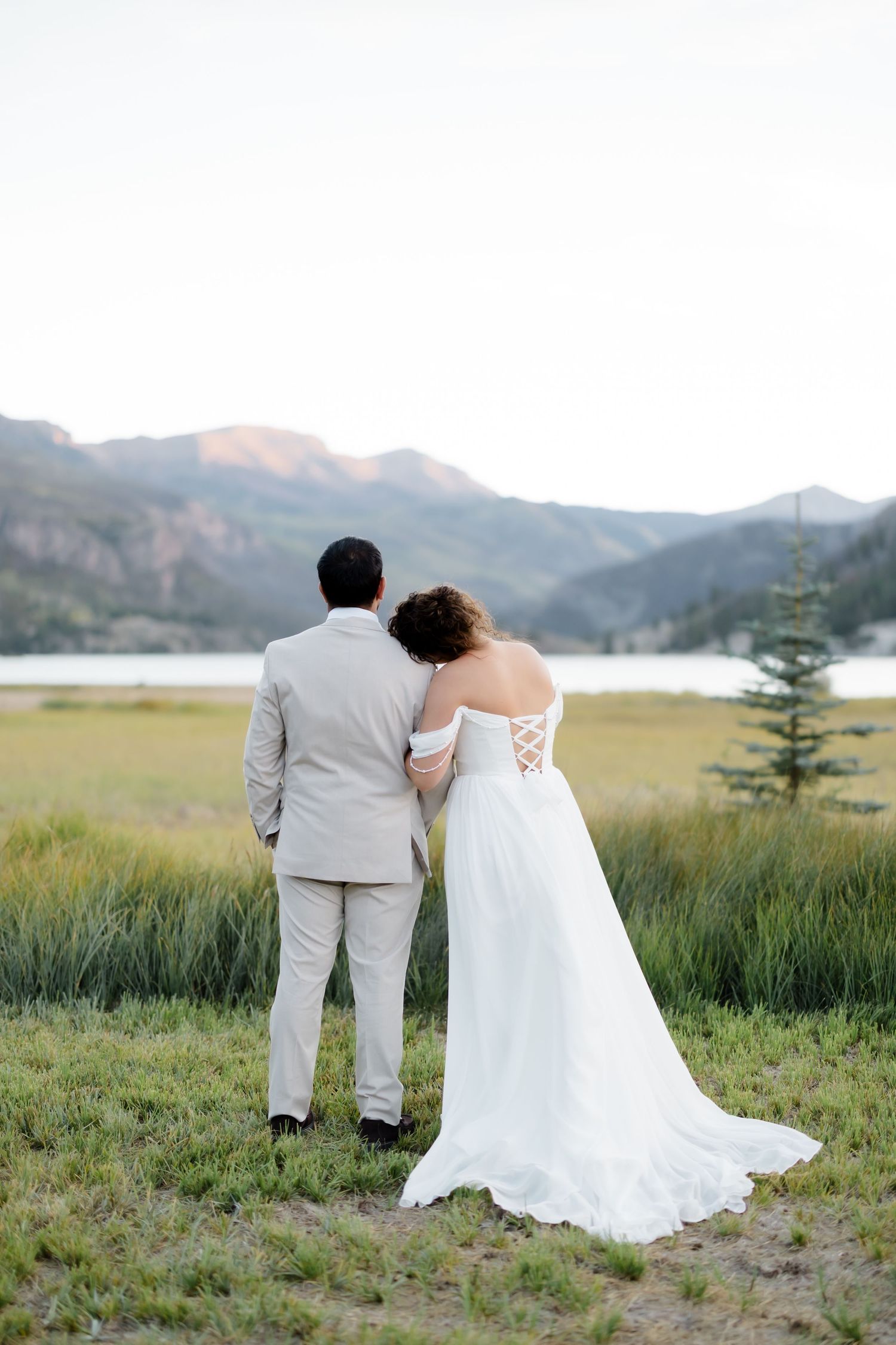 Bride and groom stand arm in arm facing a scenic mountain lake surrounded by lush green meadows.