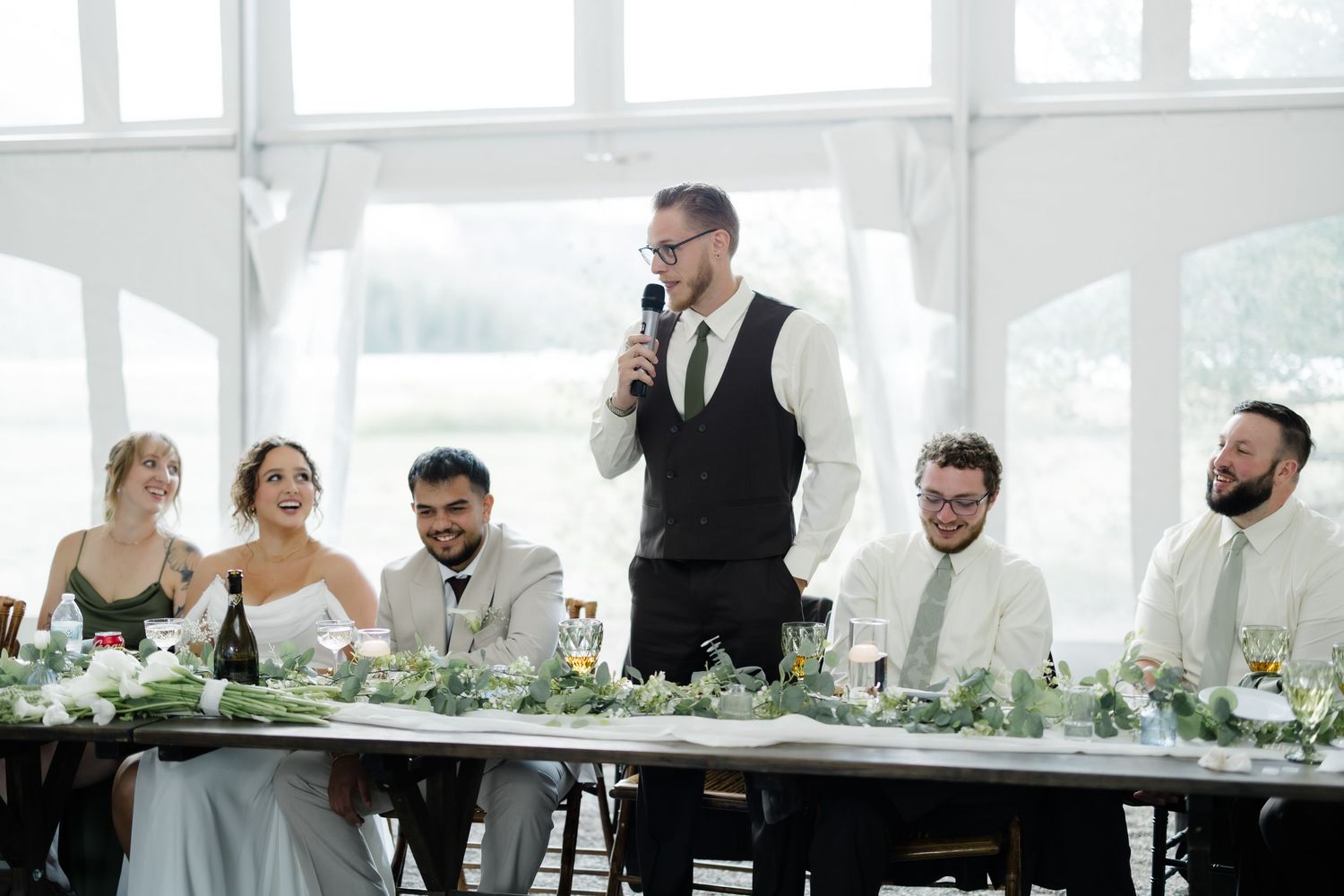 Best man delivers a heartfelt speech at wedding head table decorated with greenery and white flowers.