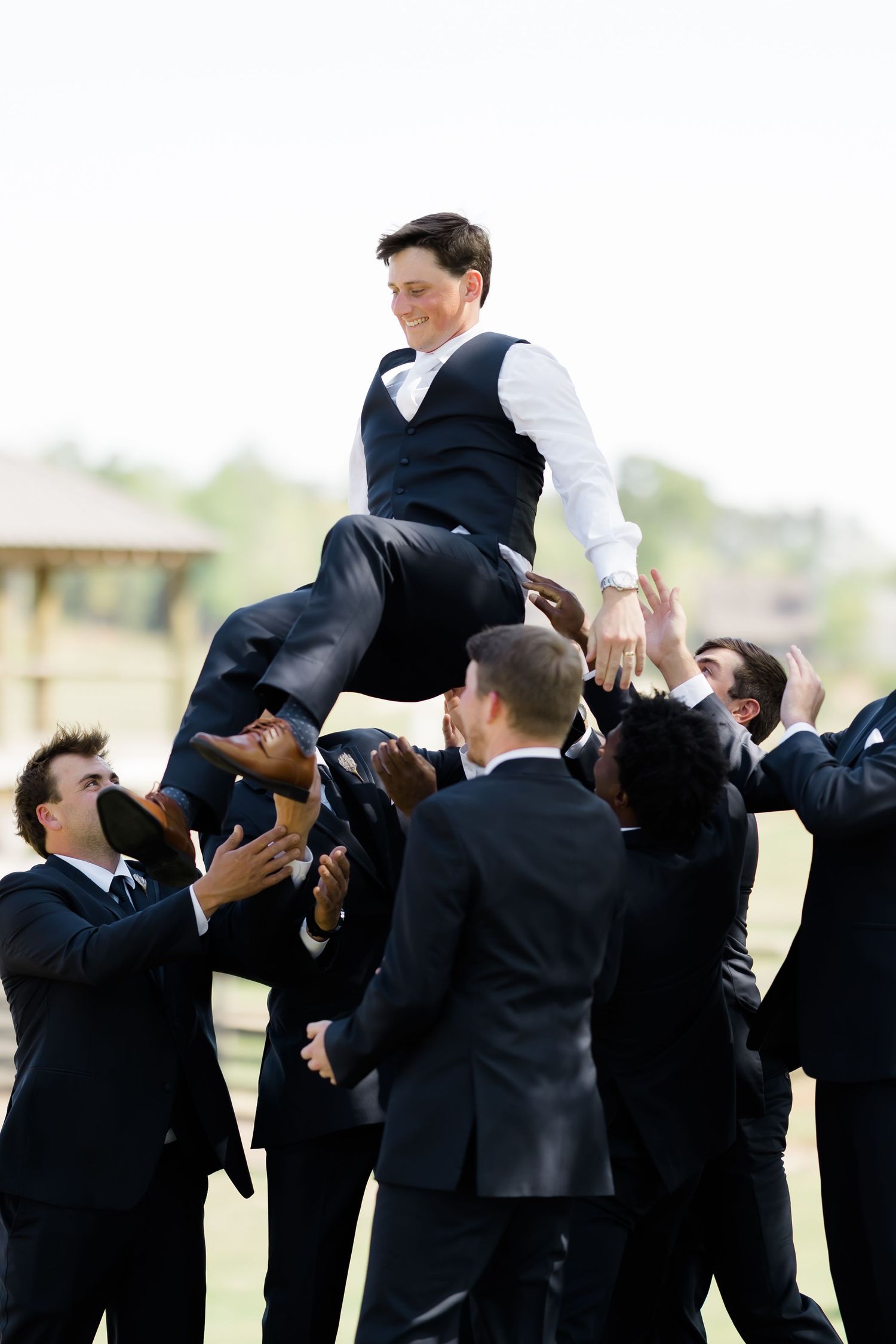 Groomsmen tossing the groom into the air outdoors at a wedding celebration.