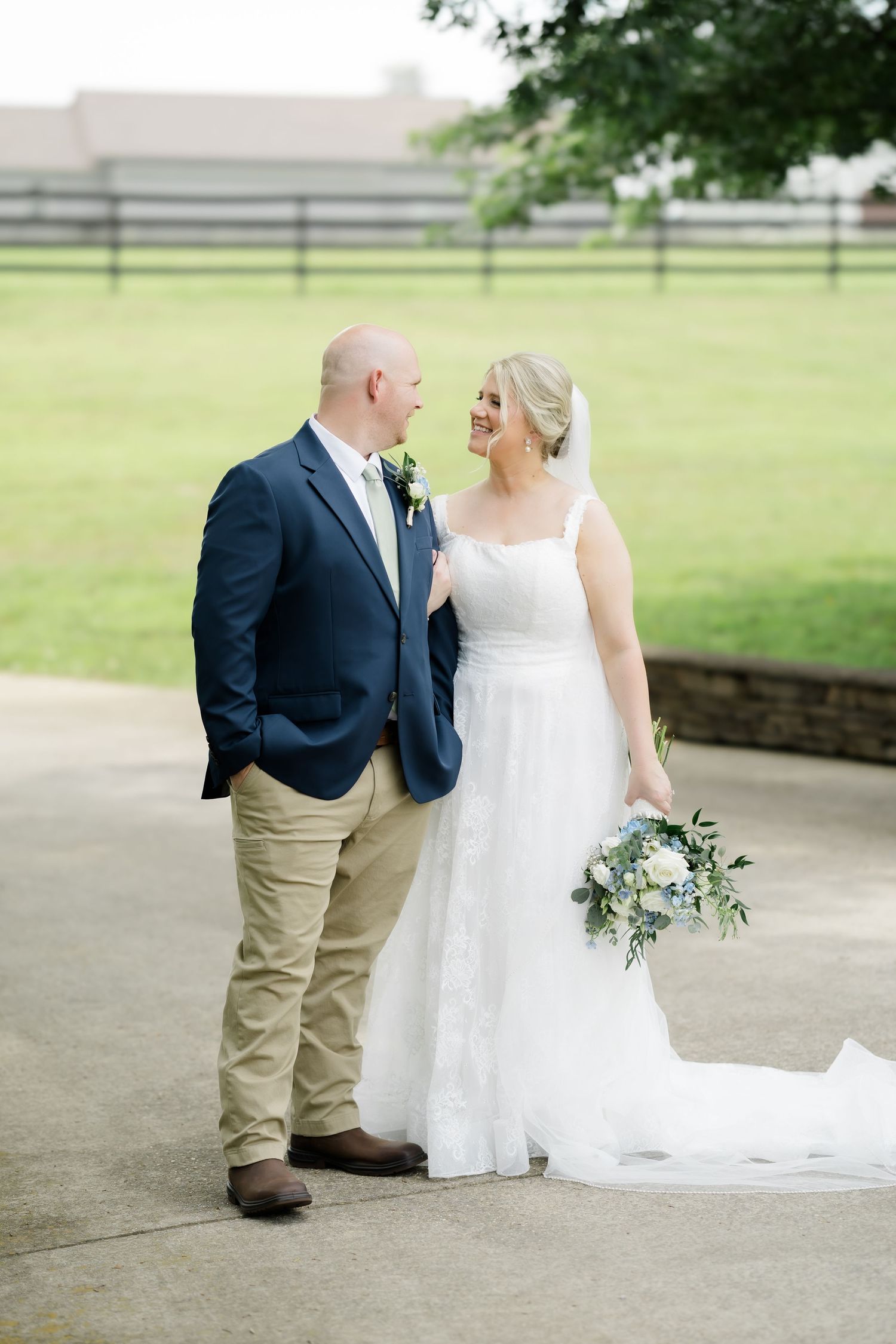 Bride and groom share a loving glance outdoors, she holds a blue and white bouquet in a lace gown, he wears a navy blazer.