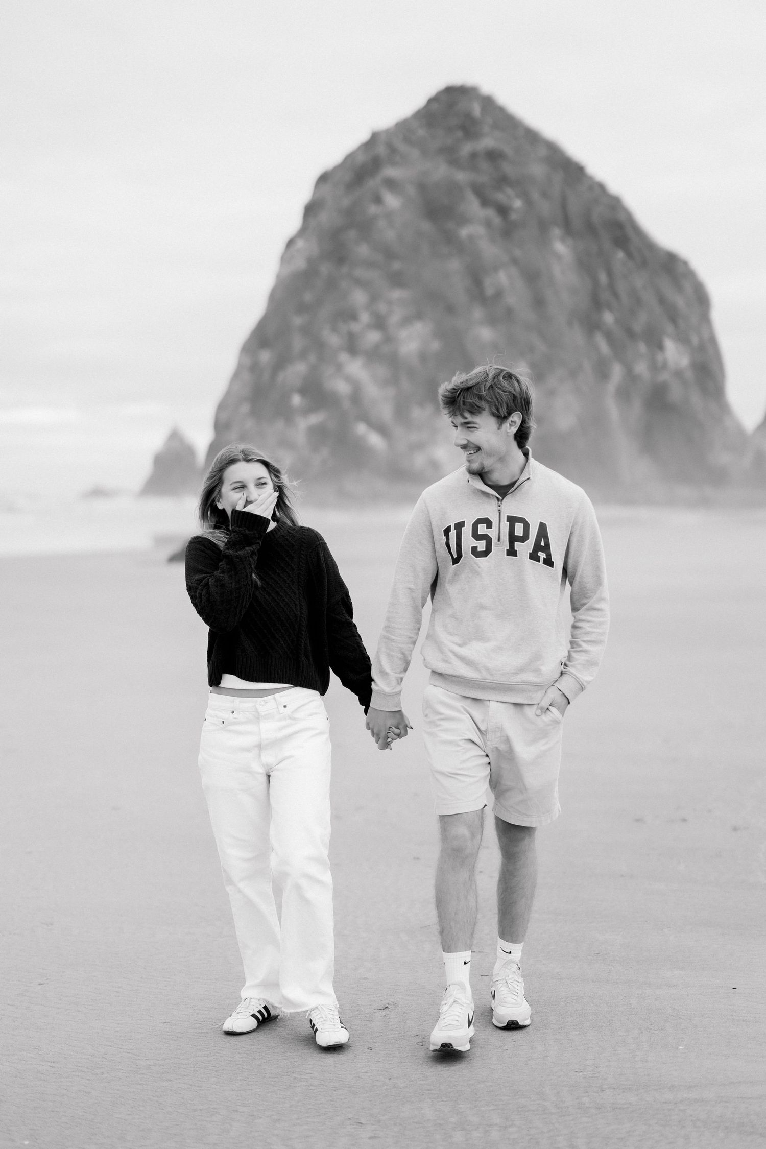 A young couple holds hands walking on a beach with Haystack Rock in the background in this black and white photo.