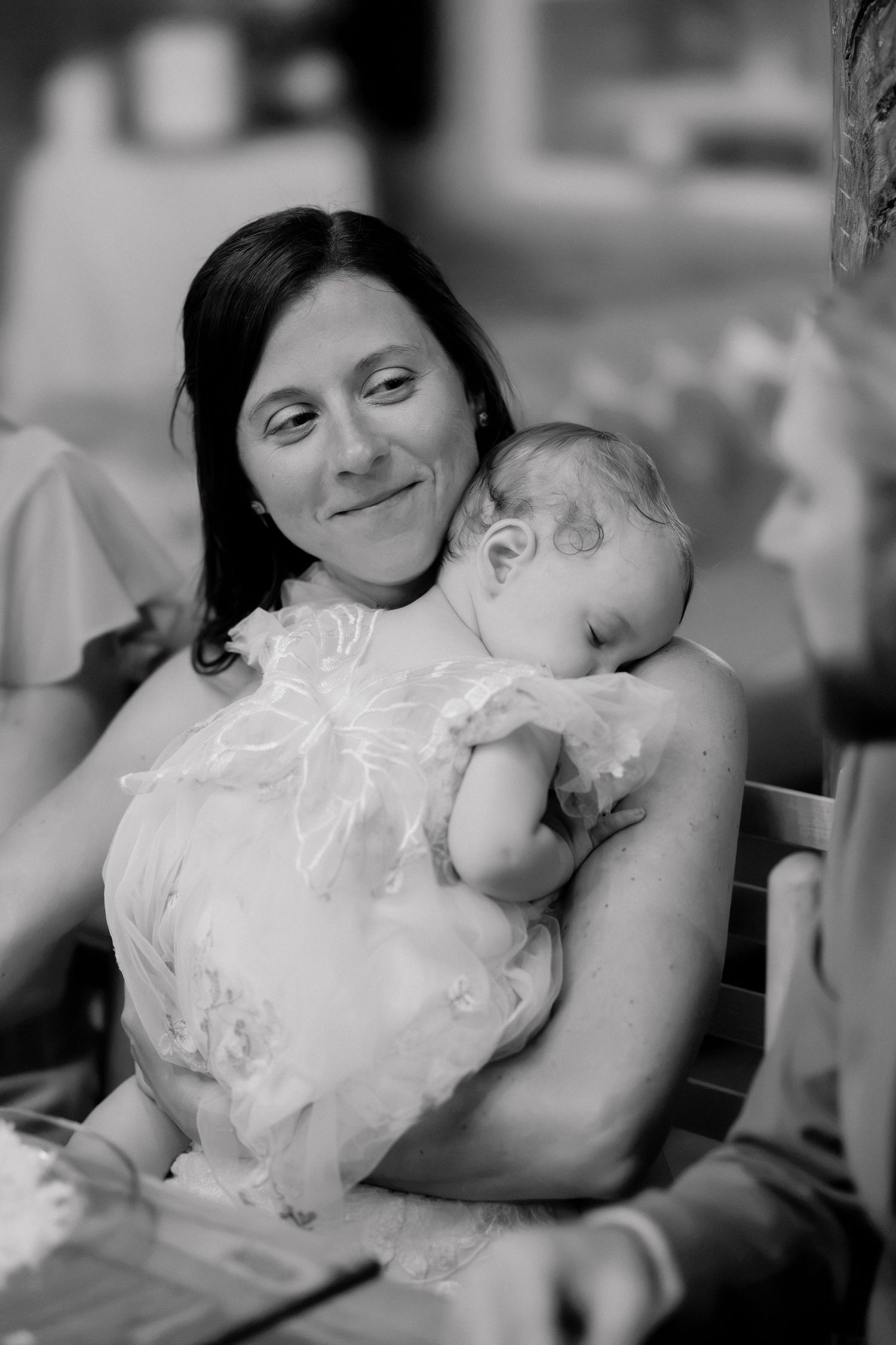 Black and white photo of a smiling woman holding a sleeping baby in a white dress at a family gathering.